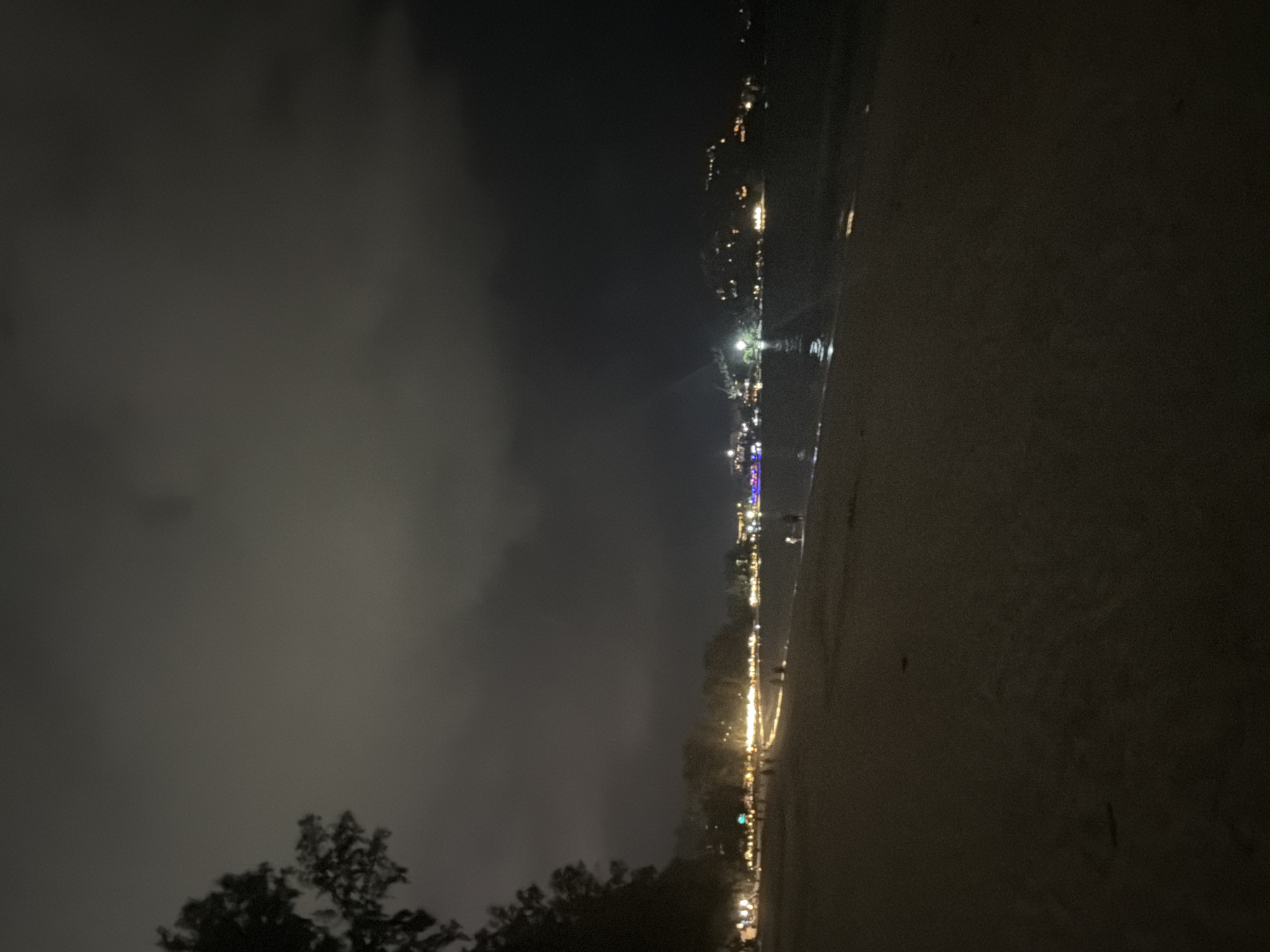 The image shows a nighttime view of a beach in Choeng Thale, Thailand. The beach is dark, with a wet sand surface reflecting some of the lights from the buildings and streetlights along the coastline. The sky is overcast with heavy clouds. A line of lights, likely from buildings and streetlights, stretches along the distant coastline. Some trees can be seen in the foreground, silhouetted against the night sky. There are no visible people or specific actions taking place.