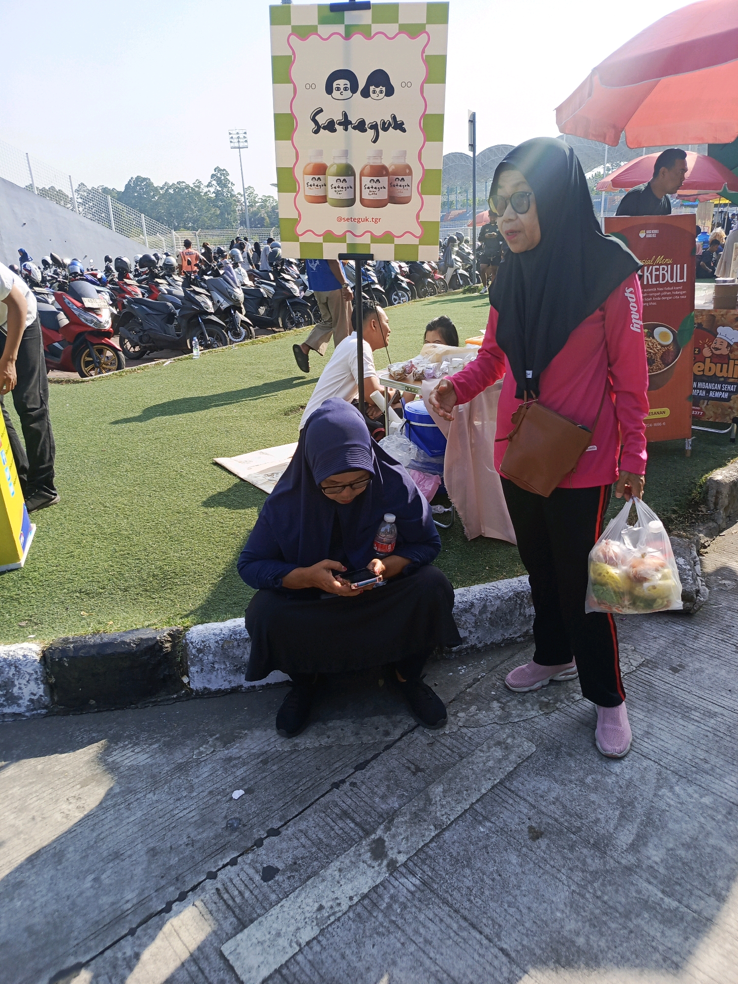 In an outdoor setting, likely a market or event space in Tangerang, Indonesia, two women are visible. One woman, wearing a dark blue hijab and glasses, is crouched down and looking at her mobile phone. She is dressed in a dark blue outer garment and dark pants. Next to her, a woman in a black hijab and a pink long-sleeved shirt is standing. She is wearing black pants with a red stripe and pink sneakers. She is holding a clear plastic bag filled with food items and a brown purse is slung across her body. In the background, a variety of motorcycles are parked, and a large banner with cartoon characters and text is displayed. The ground is a mixture of concrete and artificial turf. The scene appears to be illuminated by natural daylight.