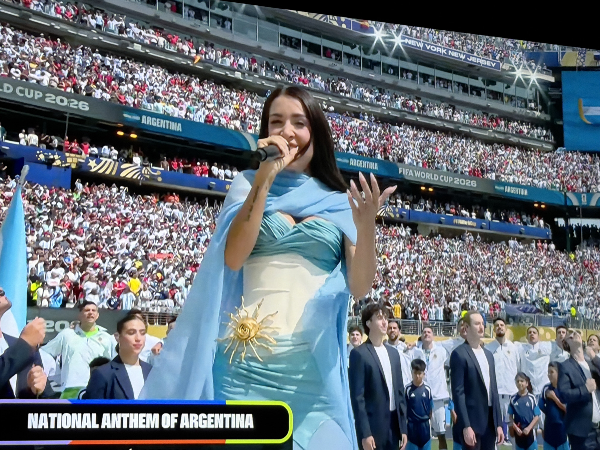 The image shows a woman singing into a microphone on a stage in a large stadium, with a crowd of people in the background. She is wearing a blue dress with a golden sun emblem on it, and a light blue scarf. The stadium has signs that read "WORLD CUP 2026" and "ARGENTINA". There are also flags visible, including one that appears to be the flag of Argentina. The scene suggests a pre-game ceremony or performance at a sporting event. The prompt requests that the location be the city of Orenburg, Russia, but there is no visual information in the image to support this specific location.
