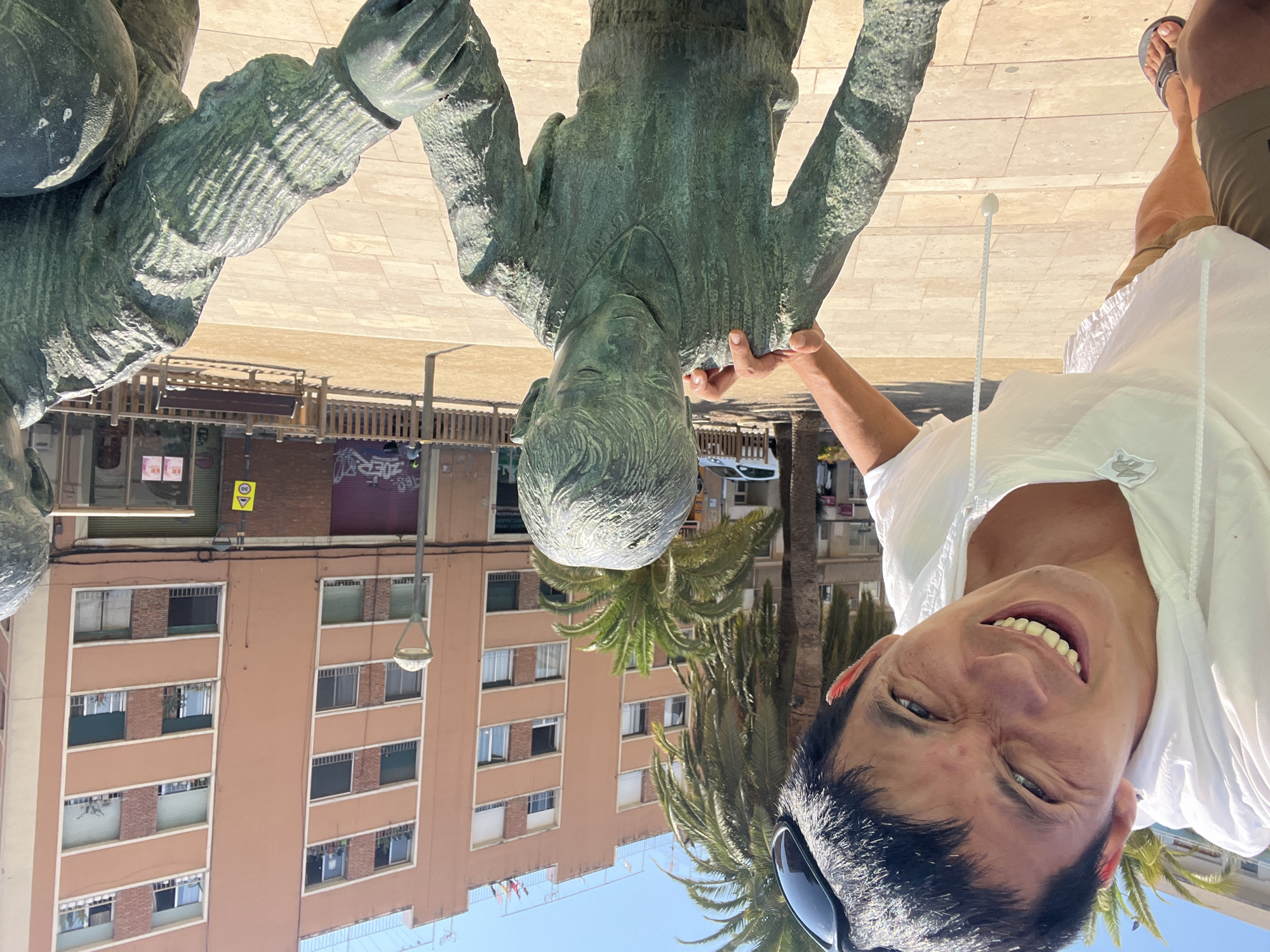 The image shows a man with a wide smile and his face contorted in a playful grimace, leaning towards the camera. He is wearing a white hooded shirt and brown shorts. His left hand is placed on the shoulder of a bronze statue of a young boy. The statue depicts the boy looking down with his hands clasped in front of him. Another bronze statue is partially visible on the right, appearing to be an adult figure. The scene is set outdoors in a paved area, with palm trees and a multi-story building in the background. The building has an orange facade with multiple windows and balconies. The presence of palm trees and the architecture suggest a Mediterranean location. This photograph was taken at Plaça dels Sedassos in Tarragona, Spain, a square known for its modern art installations and vibrant urban atmosphere. The square is a popular spot for both locals and tourists to gather and enjoy the city's ambiance.
