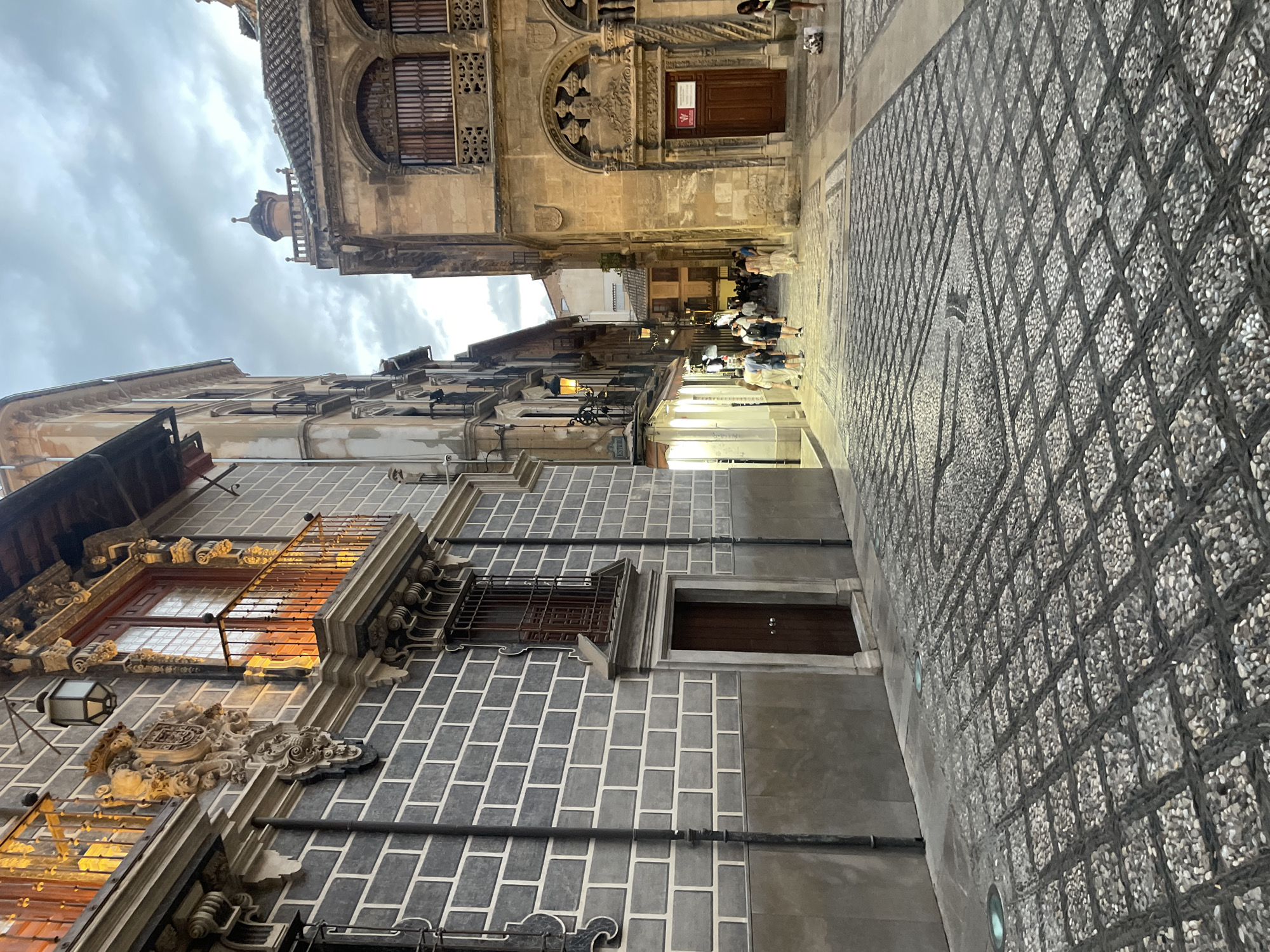 The image depicts a view looking down a narrow cobblestone street in Granada, Spain, at dusk. The street is paved with dark and light gray pebbles arranged in a diamond pattern. On the left, a building features a dark gray brick facade with white grout lines, ornate stone carvings above a second-story balcony with a wooden railing, and a dark wooden door at street level. A single lantern hangs from the facade. On the right, a building with a light brown stone facade is illuminated by interior lights, showcasing arched windows with wooden shutters and intricate decorative stonework above a large, dark wooden door. People are visible walking down the street in the distance, and the sky is overcast with a hint of twilight. The overall impression is one of a historic European street scene as evening approaches.