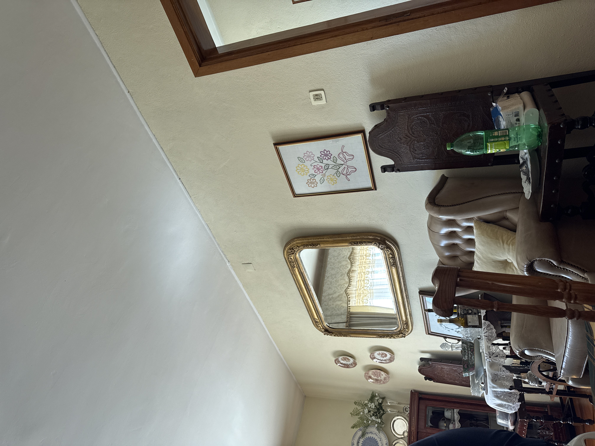 The image depicts the interior of a room. Visible are portions of a wall painted light gray, and a ceiling painted light beige. On the ceiling are a framed embroidery picture, a gold-framed mirror, and a set of decorative plates. A wooden table sits to the right of the embroidery, and a leather chair is partially visible. Furniture, including a cabinet and a table, is visible further down. A floral arrangement sits beneath the decorative plates. The city of Mirandela, Portugal, is not discernible from the image.