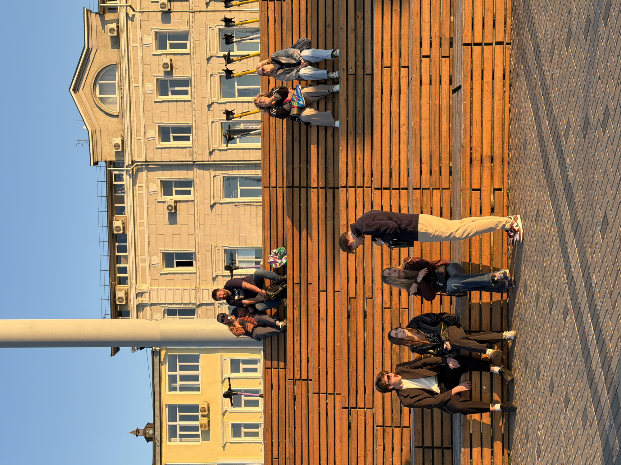 A low-angle, sun-drenched outdoor shot shows a group of young adults gathered on tiered wooden bleachers in a public square. In the background, two multi-story buildings with ornate architectural details stand under a clear blue sky. The building on the left is yellow, while the larger one on the right is a neutral stone color with several windows and air conditioning units. A tall, cylindrical concrete pillar is prominent in the left portion of the background, partially obscuring the yellow building. A line of yellow electric scooters is parked in front of the buildings.
On the upper level of the wooden bleachers, two women and two men are seated. One man is speaking, and the woman next to him is eating an ice cream cone. Another couple sits further down the row.
On a lower level of the bleachers, a group of four individuals is more centrally featured. A young man in a dark suit and white shirt is seated on the far left, with his mouth open as if speaking or singing. Next to him, two young women are seated, one in a black jacket and the other in a maroon jacket. To the right of the women, a young man in a dark sweater and light-colored pants stands, facing the seated group and appearing to be engaged in conversation. The late afternoon sun casts long shadows across the brick-patterned pavement in the foreground. This scene is located in Nizhny Novgorod, Russia, likely on the Nizhny Novgorod embankment.
