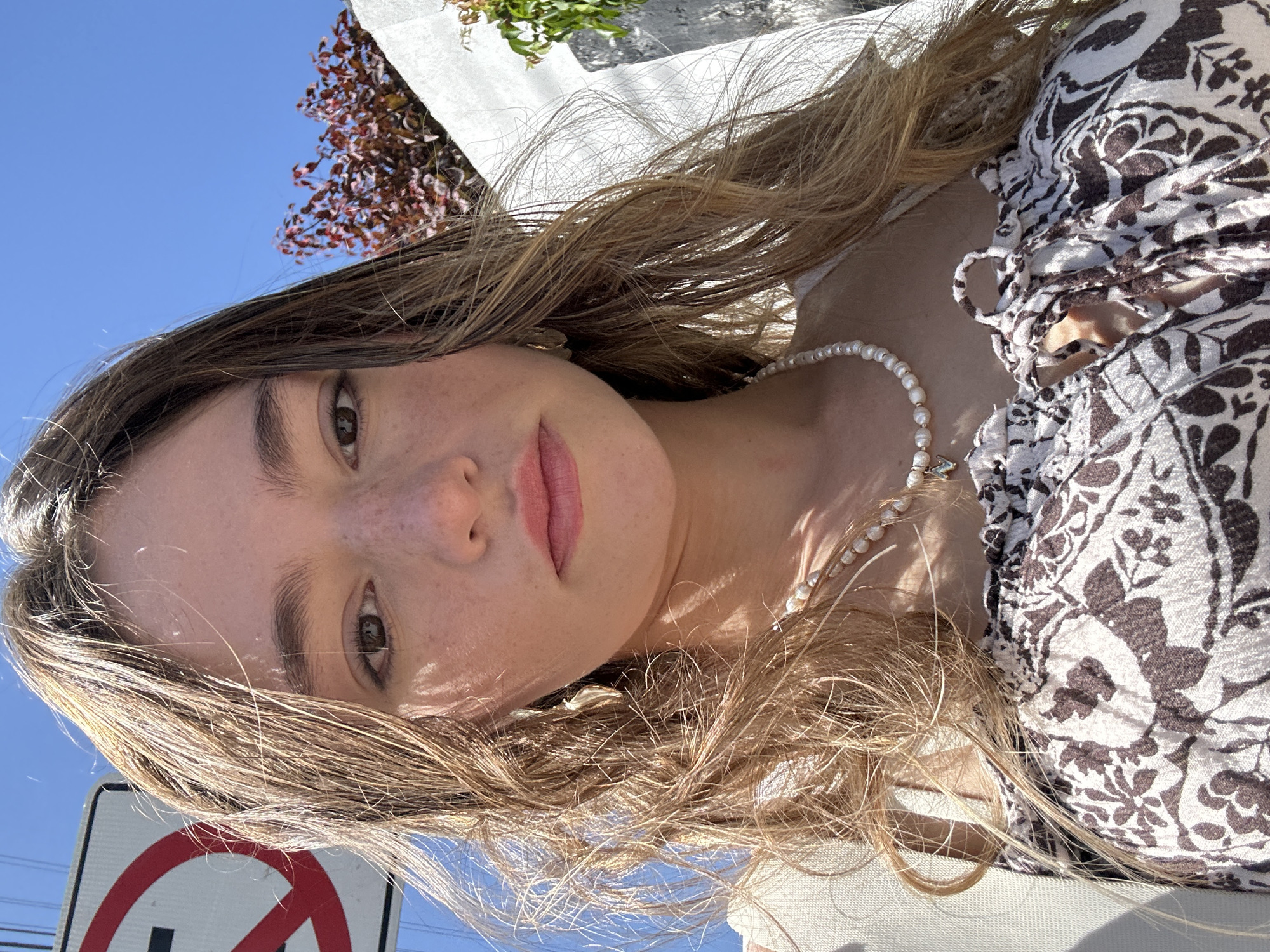 A young woman with blonde, wavy hair and light skin with freckles poses for a selfie outdoors under a bright blue sky. She is wearing a patterned off-the-shoulder top and a pearl necklace with a small pendant. Her hair is windblown and frames her face. In the background, a white wall, some reddish-brown foliage, and a portion of a white sign with a red circle and slash symbol are visible. The setting appears to be the city of San Bernardino Tlaxcalancingo, Mexico.