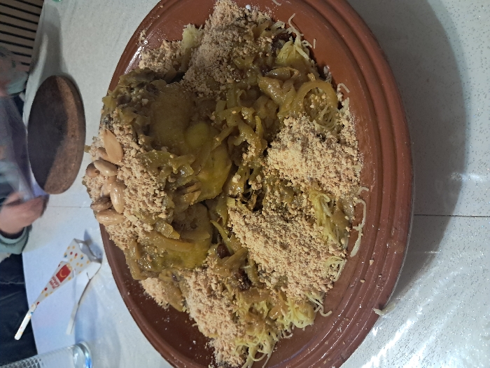 A large, brown, earthenware bowl overflows with a generous serving of Moroccan Seffa. Thin strands of noodles intertwine with a savory, golden-brown chicken stew, rich with caramelized onions and perhaps a hint of saffron. A liberal dusting of finely ground nuts blankets the dish, contrasting with scattered almonds. A decorated cone-shaped object and glasses sit on a table near the food bowl.