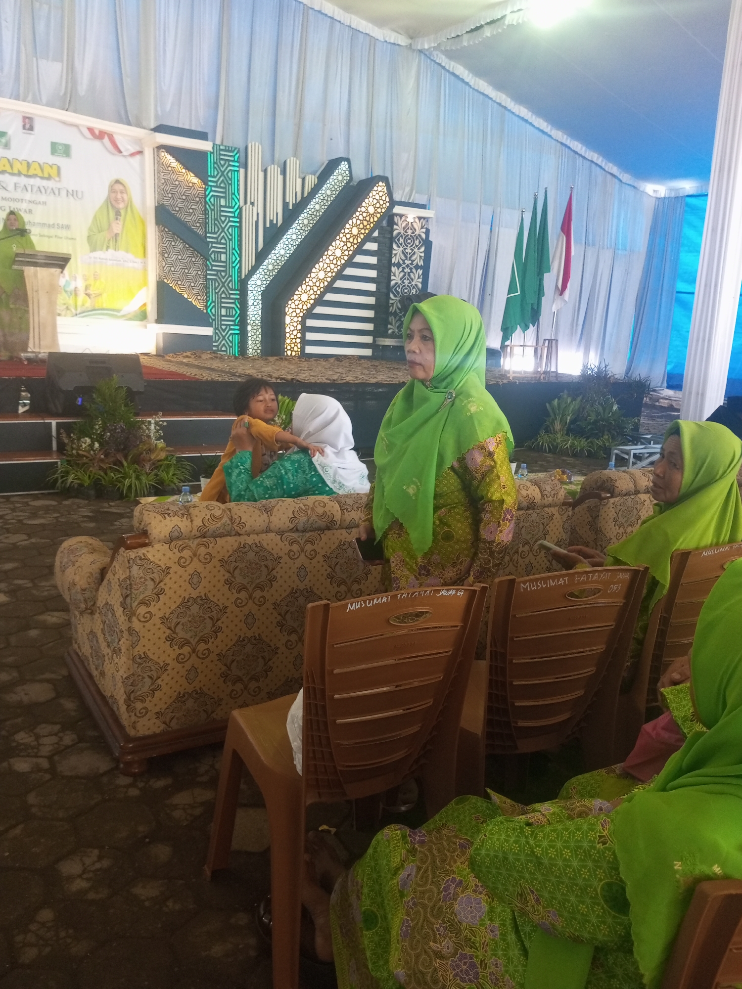 In a tented outdoor event space in Klaseman, Indonesia, several people are gathered. A woman wearing a green hijab and a patterned yellow and green outfit stands at a podium on a stage decorated with Islamic patterns and flags. To her right, other women in green hijabs are seated on couches and chairs. One woman, wearing a white hijab and a green outfit, is holding a child. The environment appears to be a communal gathering or ceremony.