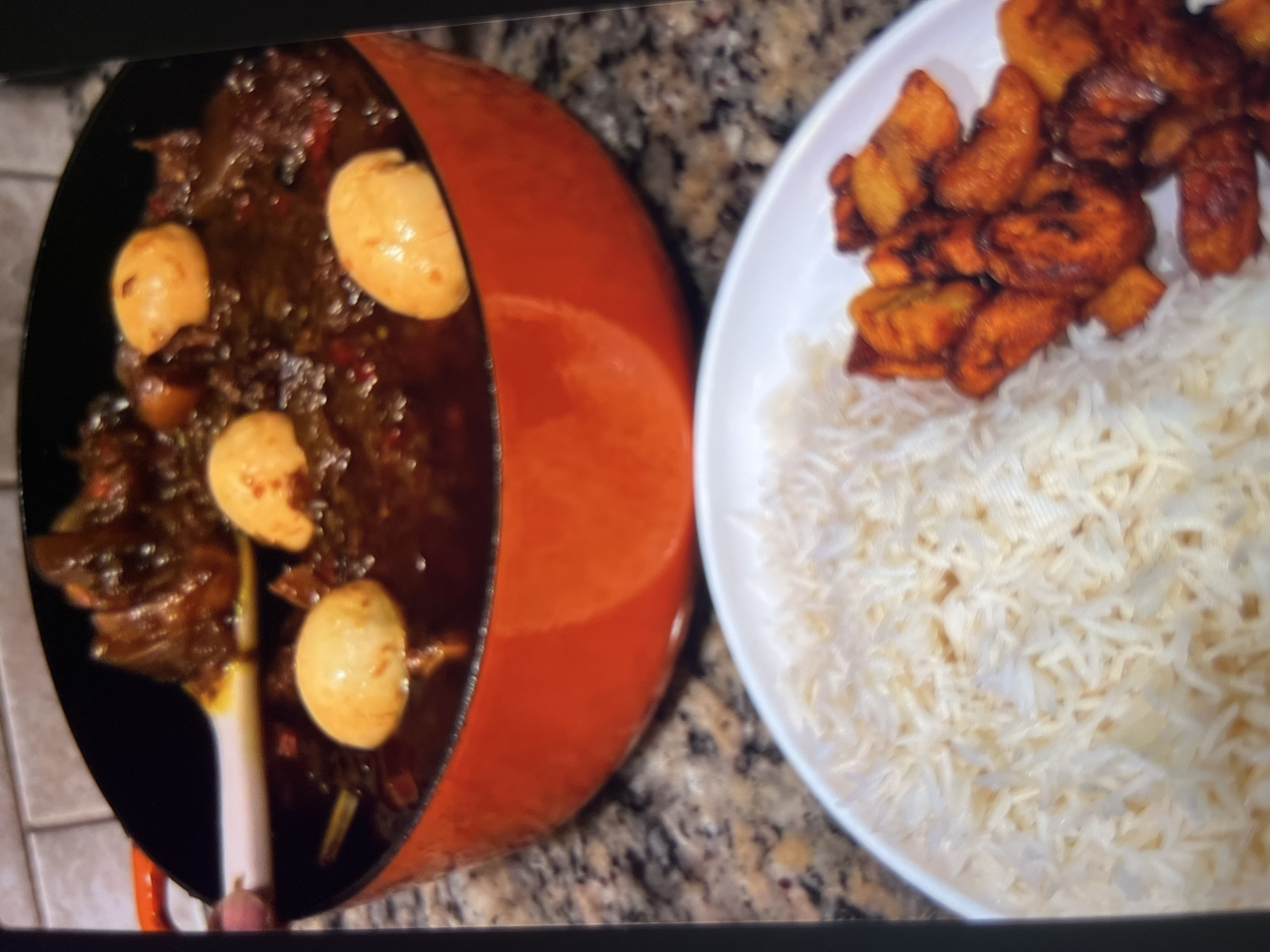 The image shows a close-up view of a meal. In the foreground, a white plate is filled with fluffy white rice on the left and fried plantains on the right. Behind the plate, a large orange pot is brimming with a dark, rich stew. Several boiled eggs are visible within the stew, along with pieces of meat and vegetables. A white spoon is lifting some of the stew from the pot, revealing its thick consistency. The background shows a speckled countertop and a tiled wall. The meal appears to be prepared in a home kitchen setting, likely in Abuja, Nigeria, given the typical ingredients and presentation of such dishes in West African cuisine.