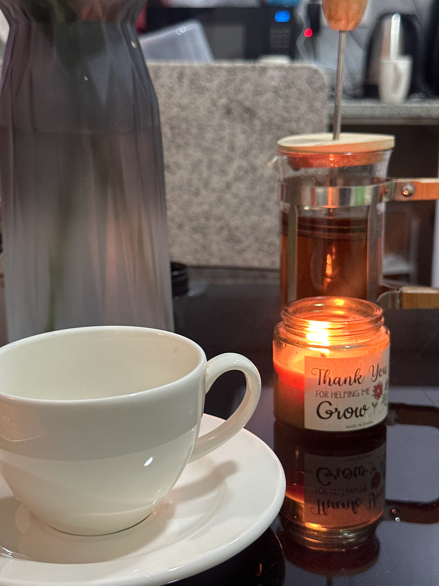 The image depicts a close-up, eye-level view of a tranquil scene, likely set in a domestic environment in Lalitpur, Nepal. In the foreground, a white ceramic teacup and saucer rest on a reflective black surface. To the right of the teacup, a small glass candle jar glows warmly, its flame illuminating a label that reads "Thank You for Helping Me Grow" and "Made in China." Behind the teacup and candle, a glass French press filled with amber-colored liquid steams gently, suggesting it has just been used to brew tea or coffee. A tall, dark grey vase stands to the left of the French press, its form softened by the ambient light. In the background, a textured grey surface and a hint of electronic equipment, including a blue light, are visible. The overall impression is one of calm and thoughtful relaxation.