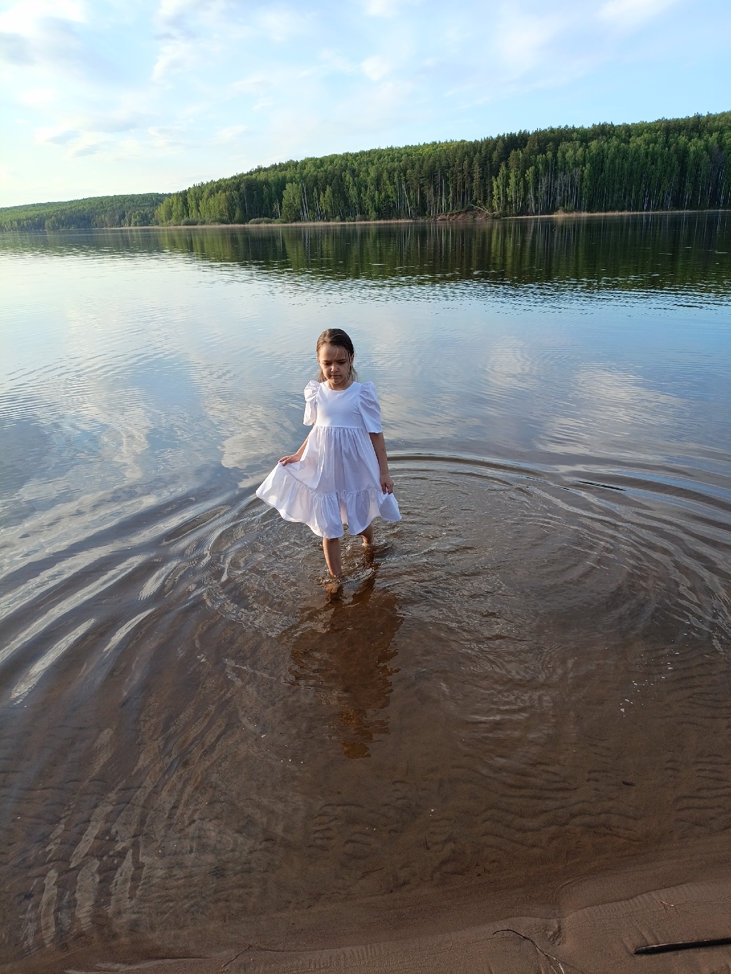 A young girl in a white dress stands in the water near a shoreline. The water is clear and reflects the sky, trees, and clouds. Ripples emanate from her feet. Behind her is a line of green trees. The setting appears to be a lake or river, possibly near Chaykovsky, Russia, given the location information. The scene is calm and peaceful.