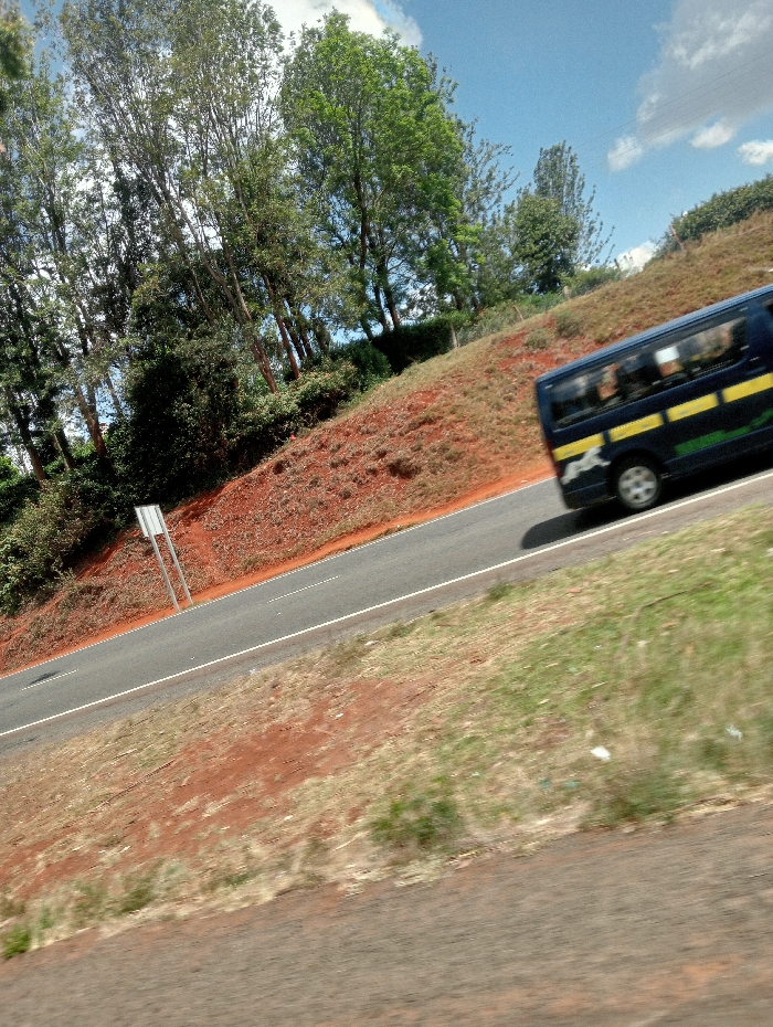 A road cuts through a landscape with a red-brown hillside covered in sparse vegetation. Trees line the hilltop on the left. A dark blue bus with a yellow stripe is moving along the road to the right. The sky is blue with scattered clouds. The foreground is out of focus with green grass. A road sign is visible on the left side of the road. The image has a slightly tilted perspective.