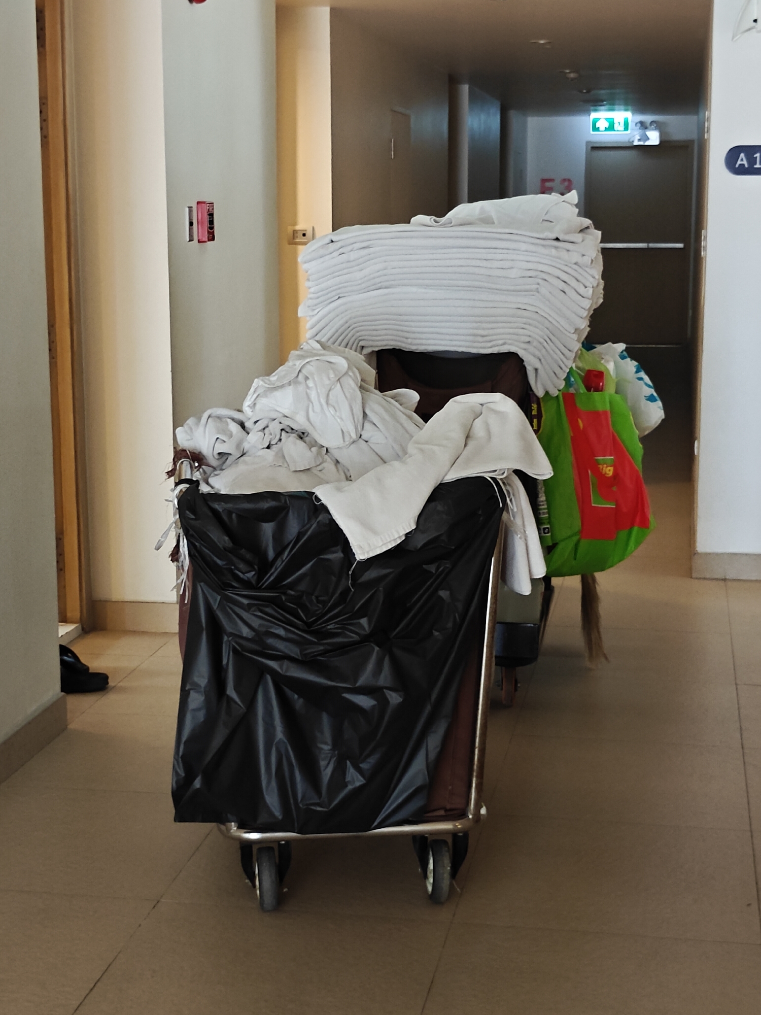 The image depicts a hallway with a laundry cart filled with white linens. The cart is positioned in the foreground, and its contents are a mixture of neatly folded and loosely draped white fabrics. A large black trash bag is attached to the front of the cart. To the right of the cart, a green bag with red accents is visible, and behind it, a white plastic bag.
The hallway itself is lit by overhead lights and an illuminated green exit sign with figures of people walking. The walls are painted in a neutral, light color. Doors are visible further down the hallway, suggesting a hotel or similar accommodation. A sign with the letter "A1" is affixed to the right wall. The floor is tiled in a beige or light brown color. A single black shoe is visible on the floor near the left edge of the image, close to a doorway. The overall impression is a service area within a building, possibly a hotel corridor, where laundry is being transported. The specific location in Pattaya City, Thailand, is not discernible from the visual elements present in the image.