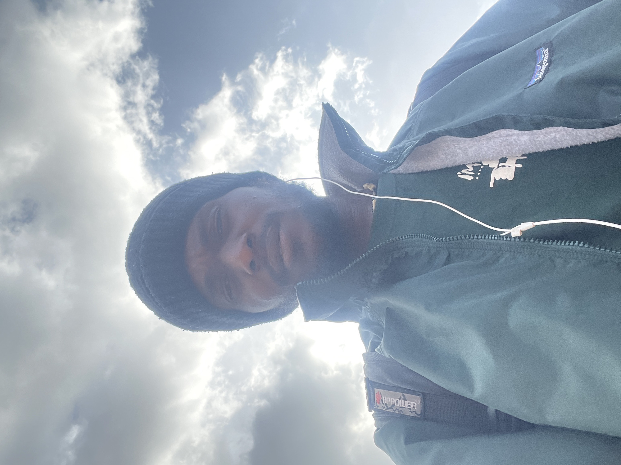 A low-angle, close-up selfie shows a Black man wearing a dark beanie, a dark green jacket zipped up over a t-shirt with white text and graphics, and white earbuds. He is looking directly at the camera, and the background is a bright, cloudy sky. The man's right shoulder reveals a dark backpack strap and a patch with camouflage print and white lettering. A Patagonia logo is visible on the right side of the jacket. The lighting suggests it is daytime. The specific location in Jos, Nigeria, cannot be determined from the image alone.