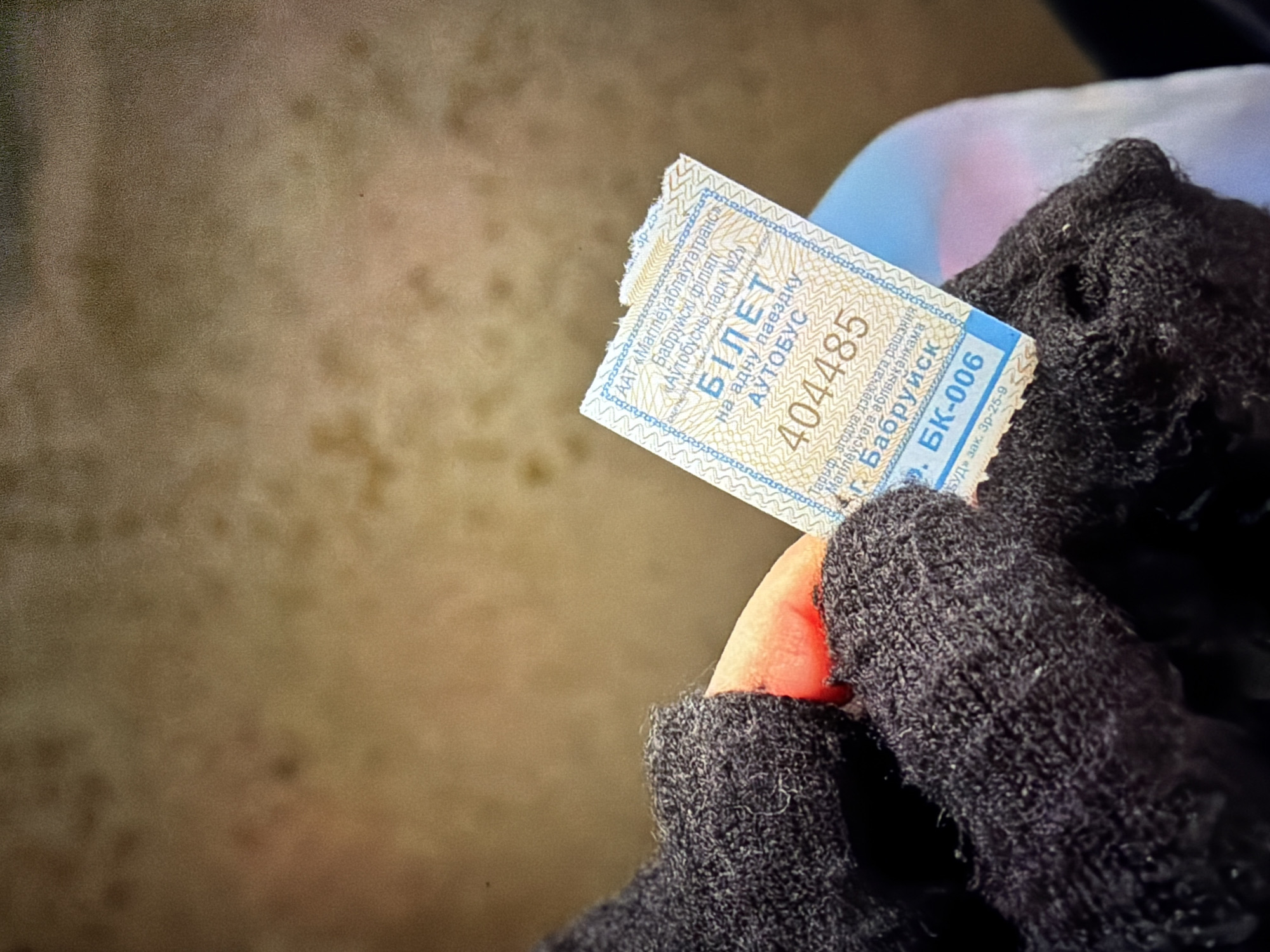 The image shows a close-up of a hand wearing a black knitted glove holding a bus ticket. The ticket is light blue and white with Cyrillic text and a serial number. The background is out of focus and appears to be a textured surface, possibly a wall or fabric. There is no indication of the city of Krasnodar, Russia.