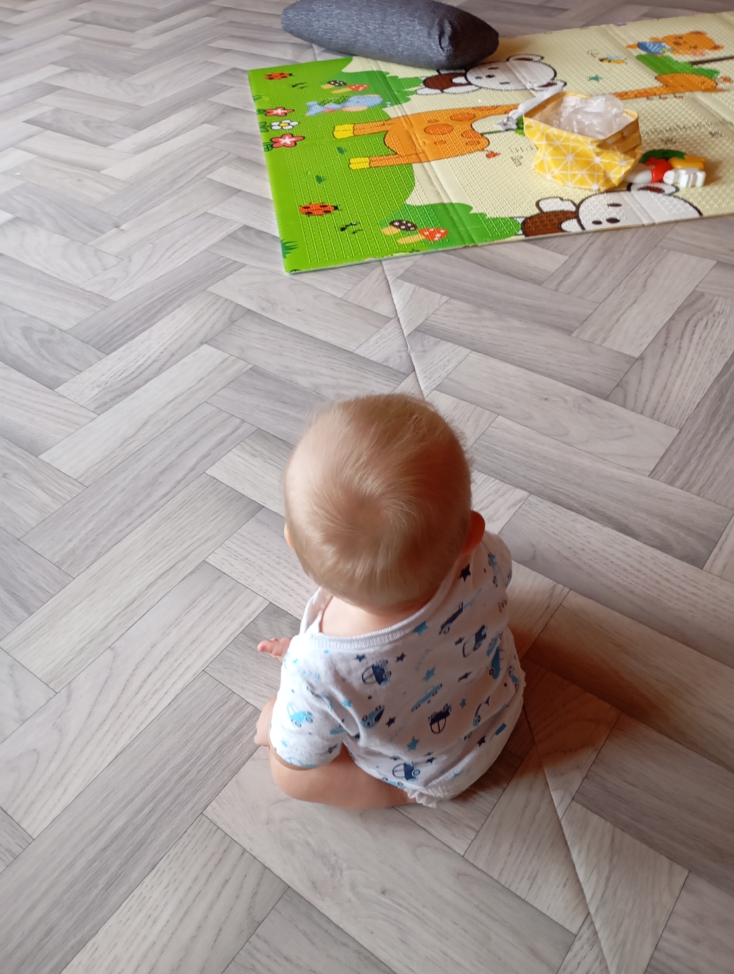 A baby is sitting on a light-colored wooden floor in an indoor setting. The baby is facing away from the camera, with their back and head visible. They are wearing a white onesie with a pattern of blue cars and stars. To the right of the baby, on the floor, is a colorful playmat with cartoon animal illustrations. On the playmat, there is a small yellow patterned bag and a few other objects, including what appears to be a toy. A gray pillow is partially visible in the upper portion of the image. The floor has a herringbone wood pattern. The scene is likely a home environment in the city of Vysokaya Gora, Russia.