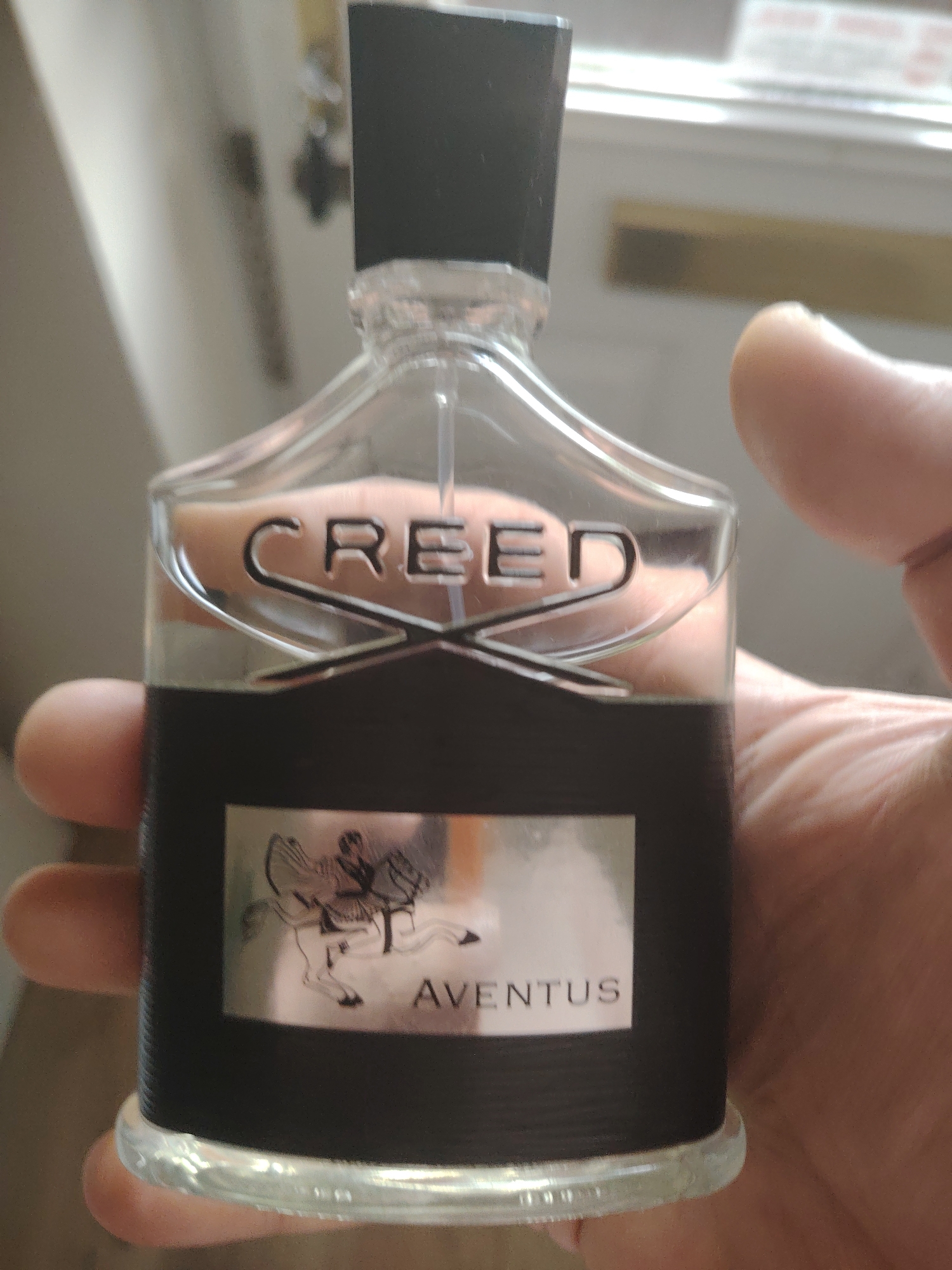The image shows a close-up of a bottle of Creed Aventus perfume, held by a person's hand. The bottle is transparent with a black cap and a black label featuring the brand name "CREED" in silver lettering. Below this, a silver label depicts a crest with a horse and rider, and the word "AVENTUS" is printed. The background is out of focus, showing what appears to be an indoor setting with a window and possibly a door. There is no discernible information in the image to suggest a specific location within Huntingdonshire, United Kingdom.