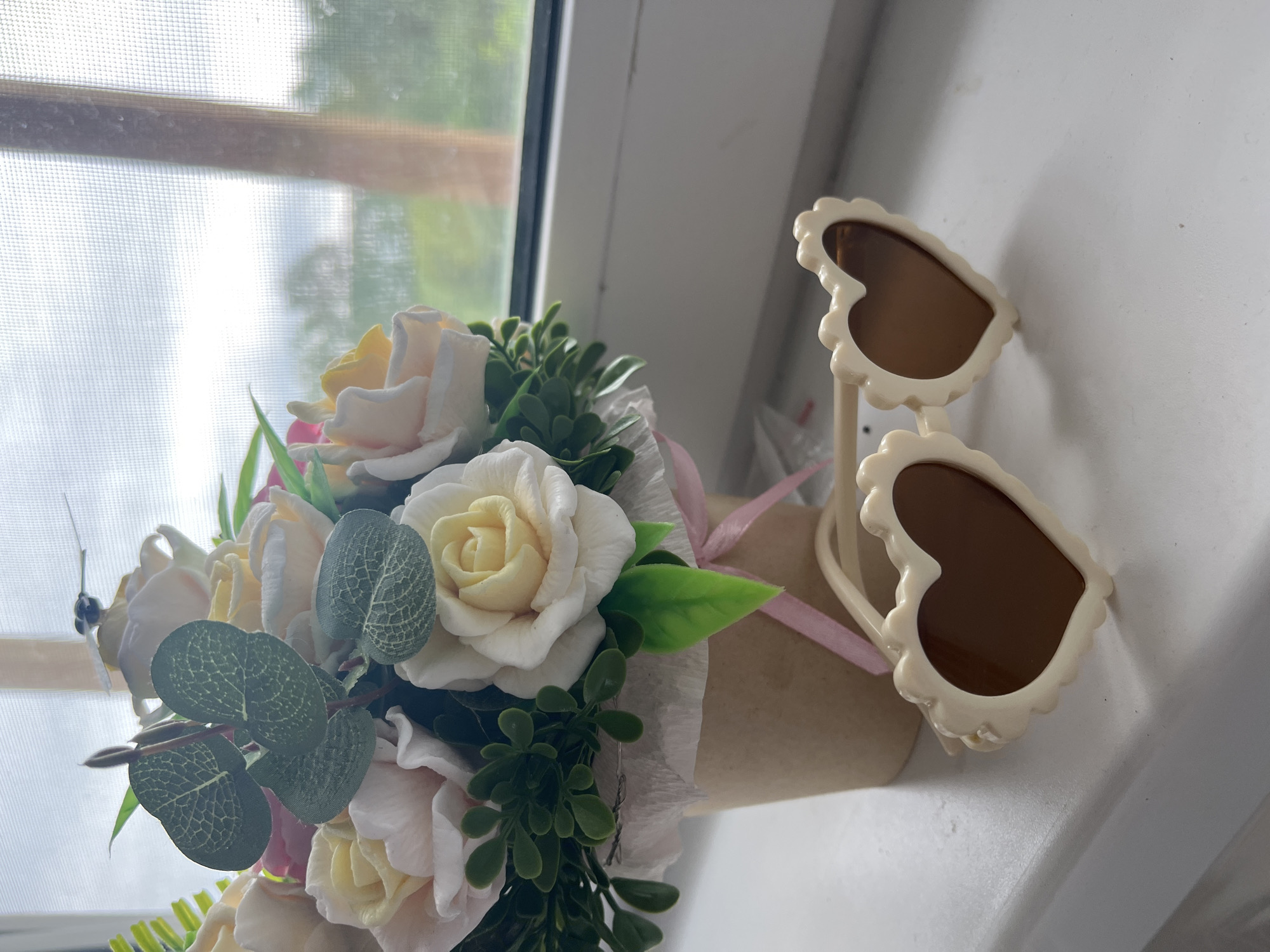 The image depicts a still life arrangement on a white surface, likely a windowsill or counter. A bouquet of artificial roses, mostly in white and pale yellow hues, is presented in a brown paper cone, tied with a pale pink ribbon. The roses are interspersed with green foliage. To the right of the bouquet, a pair of heart-shaped sunglasses with a light beige frame and brown lenses rests on the surface. The background includes a window covered with a screen, allowing a view of greenery outside. The location is Unknown, Unknown.