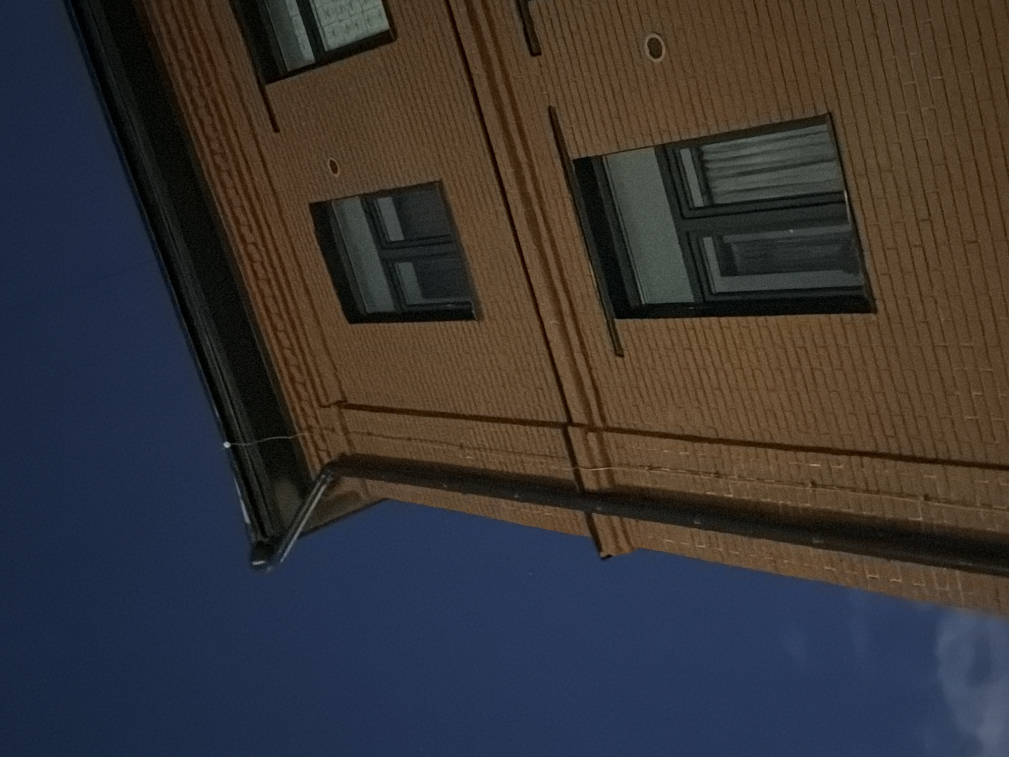 A brick building with dark windows stands against a dark blue sky. The building has a black gutter running along the roofline. One window on the upper floor is partially visible, and below it is a larger window with curtains. The brickwork is a warm brown color, and there are decorative horizontal bands above the upper window. A dark downspout runs vertically along the side of the building. The image was taken at night in Moscow, Russia.