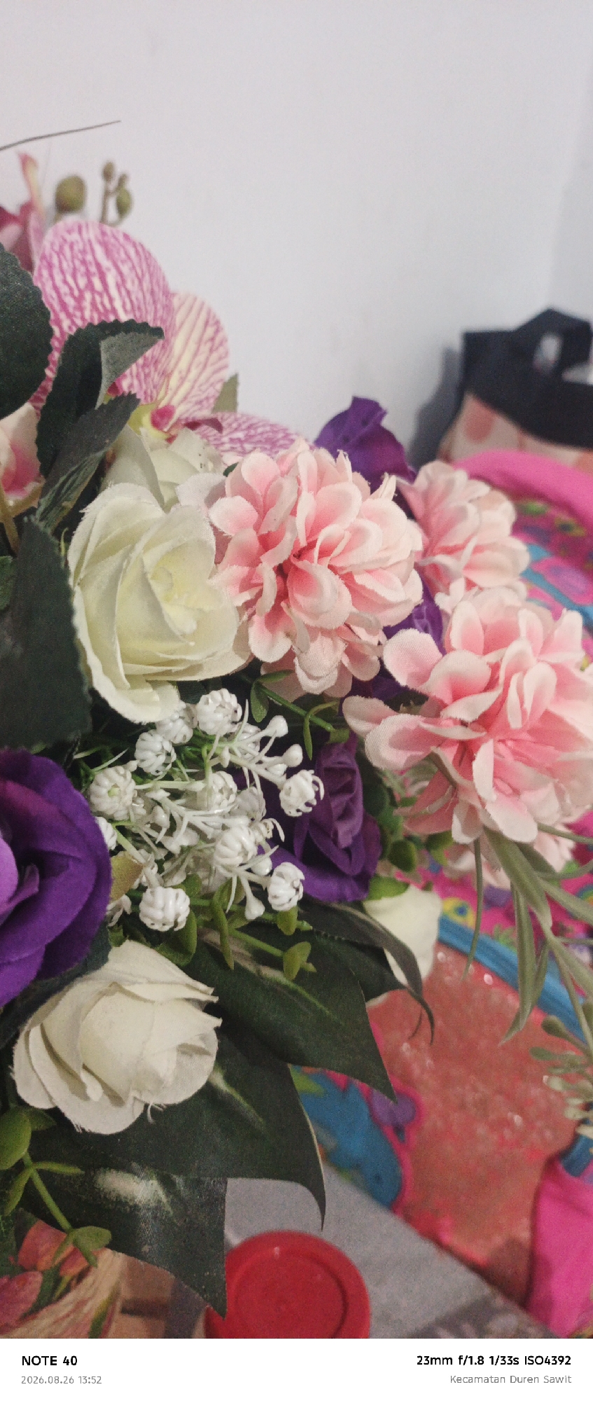 A close-up, vertical, eye-level shot showcases a vibrant artificial flower arrangement, predominantly featuring pink and white roses and orchids, interspersed with purple accents and small white filler flowers. The flowers are arranged in a bouquet, with dark green leaves providing a backdrop. In the lower right corner of the image, the text "NOTE 40" is visible, accompanied by the date and time "2026.08.26 13:52". Below this, "23mm f/1.8 1/33s ISO4392" and "Kecamatan Duren Sawit" are displayed, indicating the camera settings and the location in East Jakarta, Indonesia. The background is blurred and features a mix of colors, possibly indicating other objects or fabrics. A bright red circular object is partially visible in the bottom right corner.