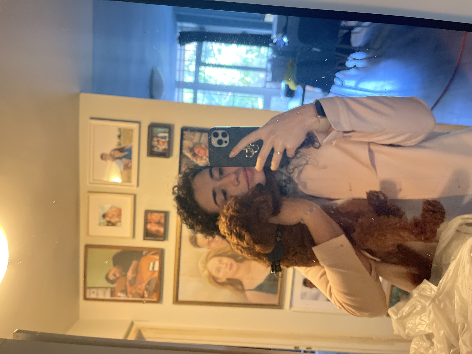 A person is taking a selfie in a mirror, holding a brown dog. The background features a wall with several framed pictures, and a window showing what appears to be an outdoor urban scene. The location is not discernible from the visual information provided to be in Sofia, Bulgaria.