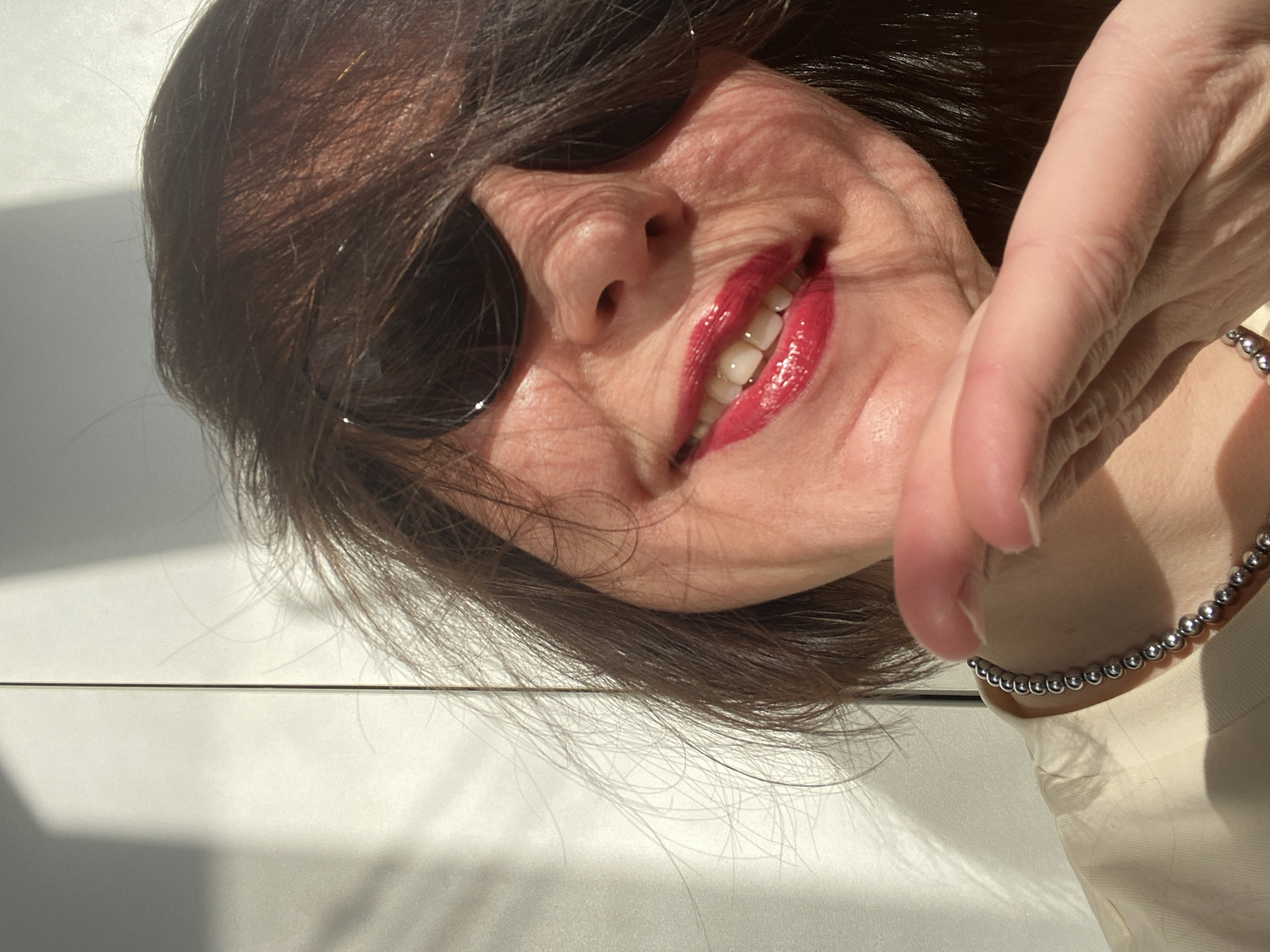 A middle-aged woman with dark hair and sunglasses smiles at the camera. Her hand, adorned with a silver beaded bracelet, is partially in the frame. She wears bright red lipstick. The background appears to be a light-colored surface, possibly a wall or table. The photo does not contain sufficient information to confirm a specific location like the city of Chaikovsky, Russia.