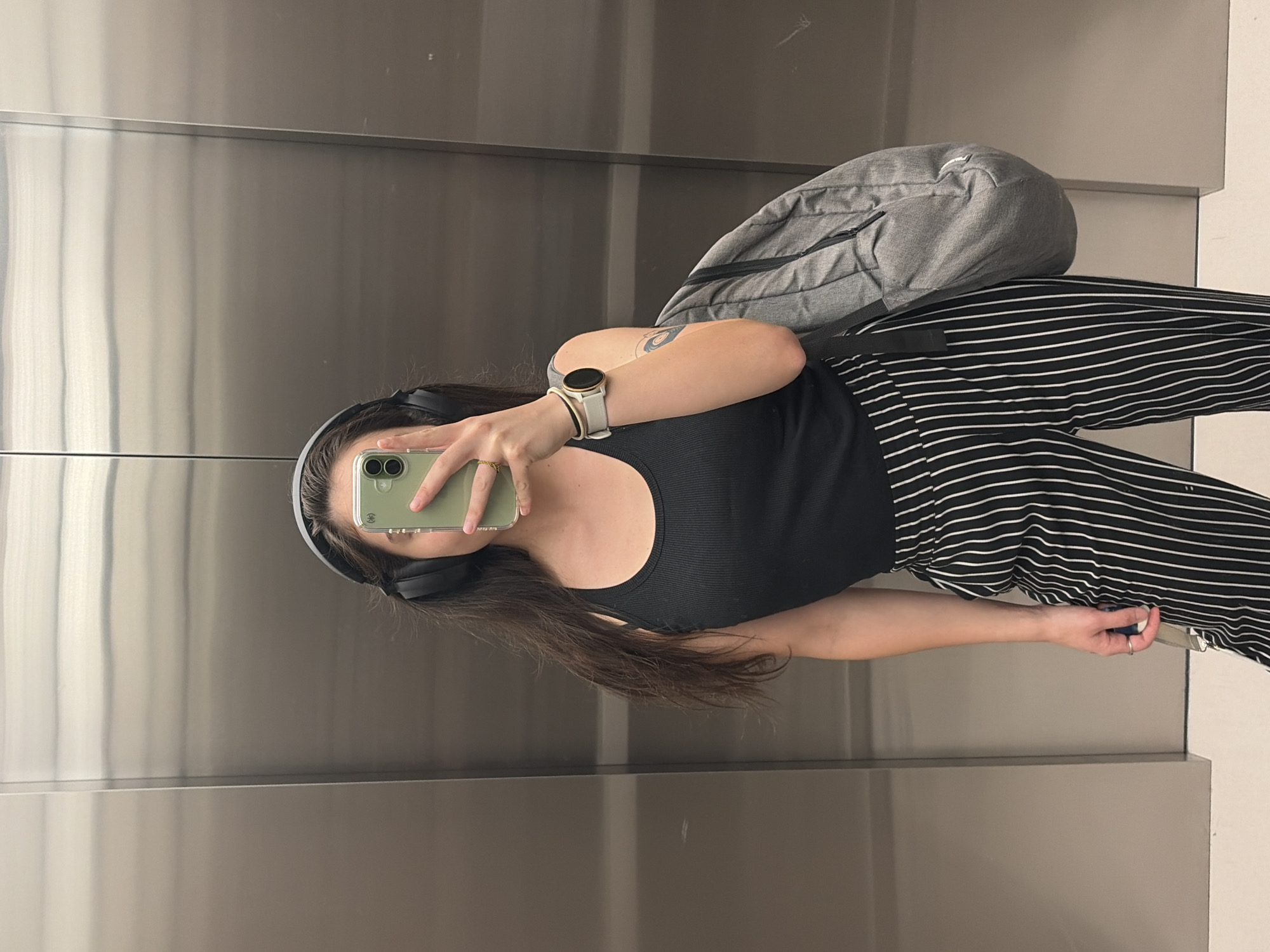 In an elevator, a person stands facing a reflective surface, taking a mirror selfie. They are wearing black headphones, a black ribbed tank top, and black pants with white vertical pinstripes. A gray backpack is slung over their right shoulder. The person's face is obscured by a light green smartphone. On their left wrist, they wear a white smartwatch with a black circular face, and a thin black bracelet. A ring is visible on their right ring finger. The elevator walls appear to be brushed metal.