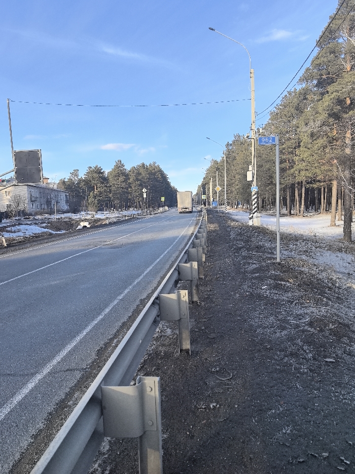 This forensic description details a roadside scene in Ablatuysky Bor, Russia.
**Location:** The scene is situated along a two-lane asphalt road identified by a blue sign as "962 km / Автодорога 22" (Highway 22 at kilometer 962).
**Time and Environment:** The image depicts a bright, clear daytime with a predominantly blue sky and faint wispy clouds. The presence of sparse snow patches on the ground suggests a late autumn or early winter season.
**Roadway and Infrastructure:**
* In the foreground, a galvanized metal guardrail runs along the right edge of the asphalt road, extending into the midground.
* The road surface is gray asphalt with a solid white edge line and a dashed white lane divider visible on the left side of the guardrail. Some minor road surface imperfections are present.
* To the immediate right of the guardrail is a dark, rocky, and sparse dirt shoulder exhibiting scattered patches of frost or ice.
* Further along the right side, a dense stand of pine trees lines the road, with snow visible on the ground beneath them.
* Multiple utility poles with streetlights are interspersed along the treeline, supporting overhead power lines.
**Signage and Objects (Right Side):**
* A prominent blue rectangular sign with white text displaying "962 km / Автодорога 22" is affixed to a utility pole.
* Adjacent to this sign is a black and white striped pole.
* Further down the road, a yellow diamond-shaped sign with a black "X" (indicating a railway crossing without barrier) and a blue square sign with a white pedestrian icon (indicating a pedestrian crossing) are visible.
**Vehicles:**
* A white semi-trailer truck is observed traveling away from the viewer in the visible lane, its rear profile prominent.
**Left Side:**
* In the background on the left, a two-story, light-colored building (possibly residential or commercial) is visible.
* A large, dark, rectangular structure, potentially an old billboard frame or industrial equipment, is mounted on a tall pole near or on the roof of this building.
* Patches of snow are present on the ground between the road and the building.
**People:** No individuals are discernibly visible within the frame.