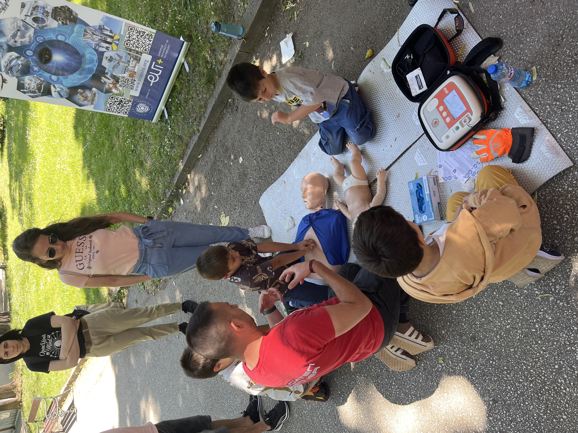 The image shows a group of people, including adults and children, gathered outdoors, likely in a park or public space. A banner with the text "EUROPEAN UNIVERSITY OF TECHNOLOGY" and the web address "www.transtu.org" is visible, along with the word "SOFIA", suggesting this is an event affiliated with the university. On a mat laid out on the ground, there is a first aid training scenario with an adult CPR manikin and an infant manikin. An adult in a red shirt is demonstrating a technique to a child. Another child is kneeling nearby, watching the AED equipment, gloves, and water bottle. Several other adults are standing around observing. One woman is wearing a "GUESS" t-shirt, and another is wearing a t-shirt with the words "Open Your Mind". The location is likely in Plovdiv, Bulgaria, inferred from the university affiliation and context. The scene depicts a first aid training or demonstration event.