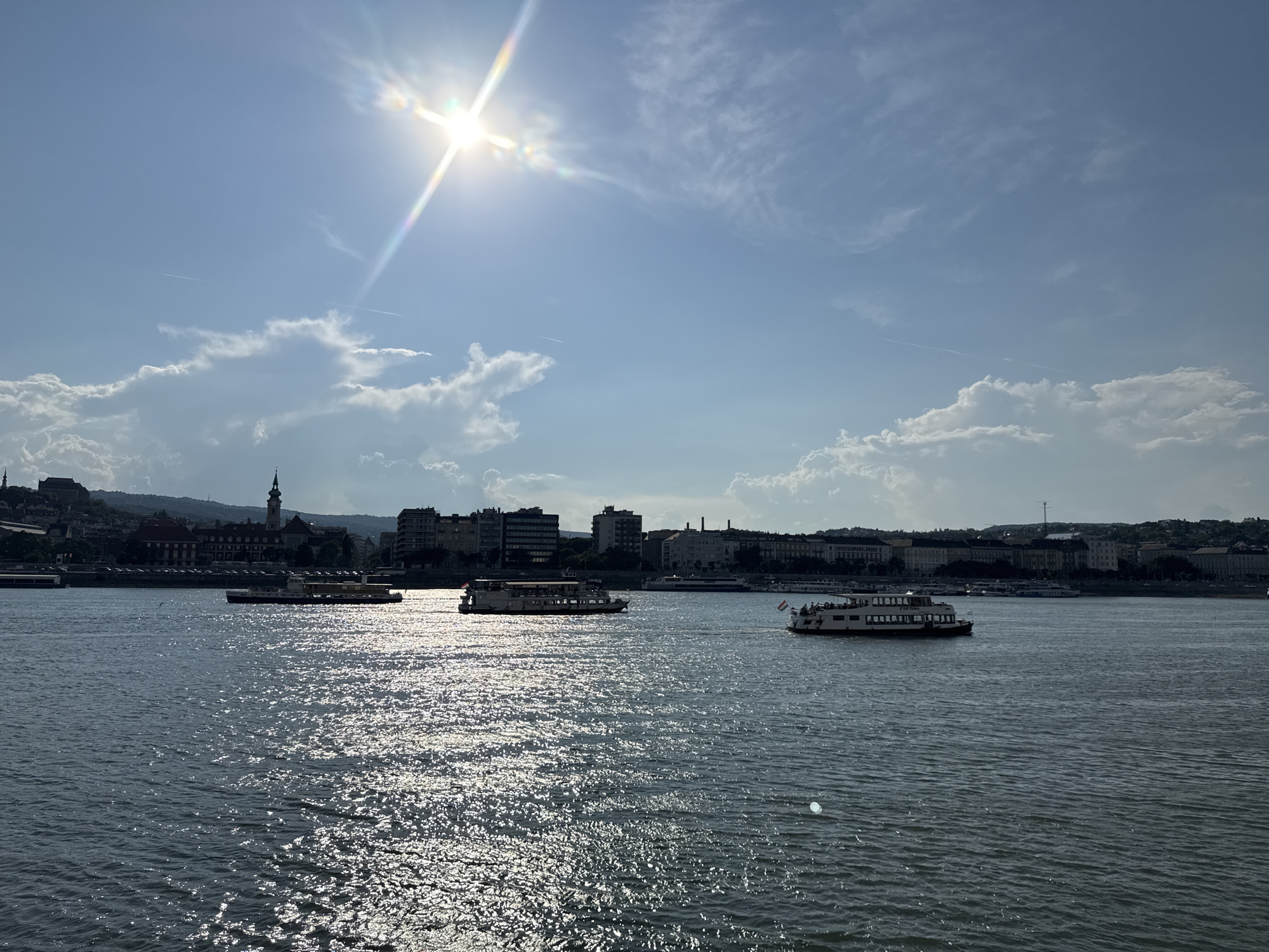 A bright, sun-drenched day unfolds over a wide river, likely the Danube in Budapest, Hungary. The sun, a brilliant starburst with visible lens flare, dominates the upper left quadrant of the sky. Wispy white clouds drift across a clear blue expanse, interspersed with larger, puffier clouds that cast subtle shadows on the water.
Several tour boats navigate the river. The closest boat on the right has passengers visible on its upper deck and displays Hungarian flags. Another boat, positioned more centrally in the frame, also carries passengers and sails towards the left. Further in the distance, more boats are visible, some appearing smaller and further away.
The river's surface is a shimmering spectacle, reflecting the bright sunlight in countless sparkling patterns. Along the riverbank, a cityscape emerges. Buildings of varying heights and architectural styles line the waterfront, with a prominent church tower visible to the left, suggesting a historic district. A hilly landscape forms the backdrop to the city. The overall impression is one of a pleasant, bustling day for tourism and river travel. The time of day appears to be mid-morning or early afternoon, given the high sun and clear conditions. No visible text is present in the image.