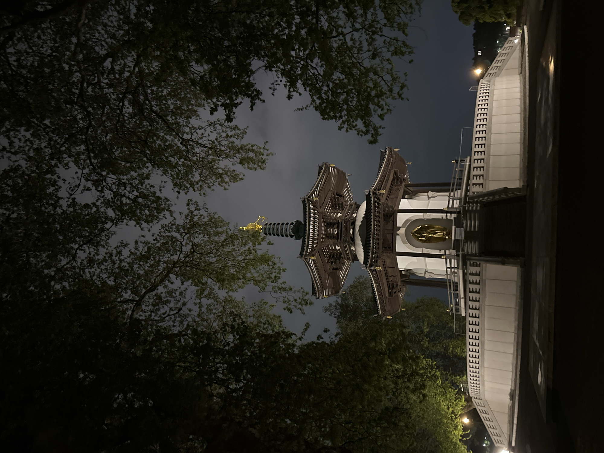 A nighttime photograph captures a multi-tiered Buddhist stupa, commonly known as a peace pagoda, framed by dark tree foliage against an overcast sky. The pagoda exhibits intricate traditional East Asian architecture with curved roofs and ornate details. A golden finial crowns the uppermost spire, and another golden figure is visible within a recessed alcove on a lower tier, both prominently lit by spotlights.
Dense, dark green trees partially obscure the structure from the left, with some foliage catching ambient light. To the right, a light-colored concrete retaining wall or walkway extends into the distance, featuring a distinctive repeating pattern of block-like shapes, resembling the letter 'E'. Distant streetlights cast an orange glow on this structure and pathway.
The dark, cloudy night sky contributes to a serene and somewhat mysterious atmosphere. The artificial lighting dramatically highlights the pagoda's features, contrasting with the deep shadows of the surrounding natural elements. No people or specific activities are visible in the scene. Given the context of London, United Kingdom, this structure is identifiable as the London Peace Pagoda located in Battersea Park.