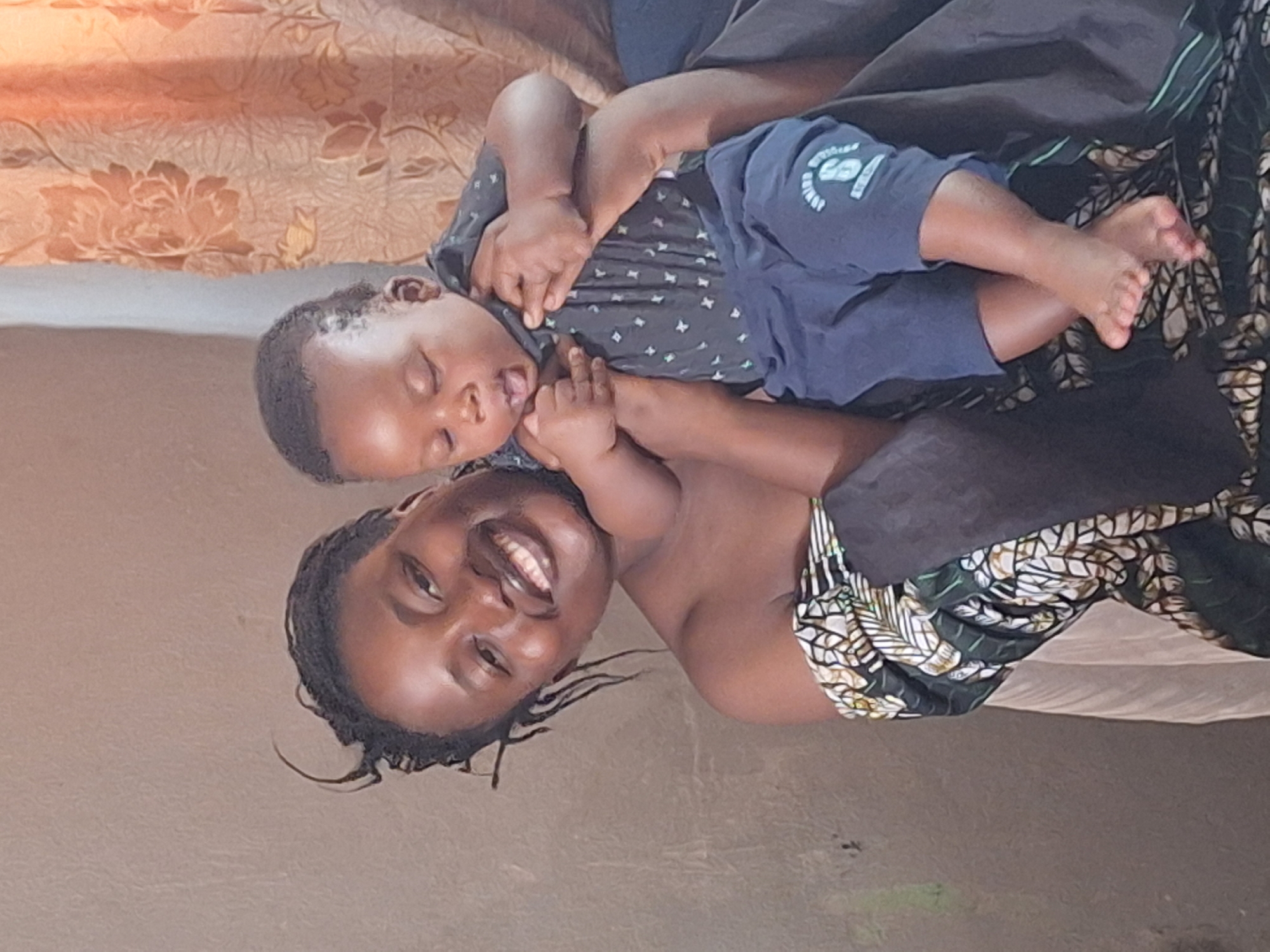 The image captures a tender moment between a woman and a baby, presented in a portrait orientation but rotated 90 degrees counter-clockwise. A woman with dark skin and dark braided hair lies on her back, looking towards the viewer with a broad, joyful smile. She is wearing a strapless garment made of a dark green or black fabric with a distinctive white and gold leaf or floral pattern.
Resting peacefully on the woman's chest and shoulder area is a sleeping baby or young toddler, also with dark skin and short dark hair. The baby's eyes are closed, and the mouth is slightly ajar, indicating deep slumber. The baby is dressed in a dark blue t-shirt adorned with a subtle, light-colored repeating pattern, and dark blue shorts or pants. On the left leg of the shorts, faint white text and a graphic are visible, including the words "BOY SQUAD" and other partially legible text like "ORIGINAL" or "EST. 2018," accompanied by a graphic resembling a head with a hat.
The background behind the woman's head reveals a light brown or peach-colored surface with a soft, embossed floral or botanical pattern, likely a bedspread or blanket. The setting appears to be indoors, under soft, warm lighting. The overall mood conveyed by the image is one of love, peace, and maternal affection.