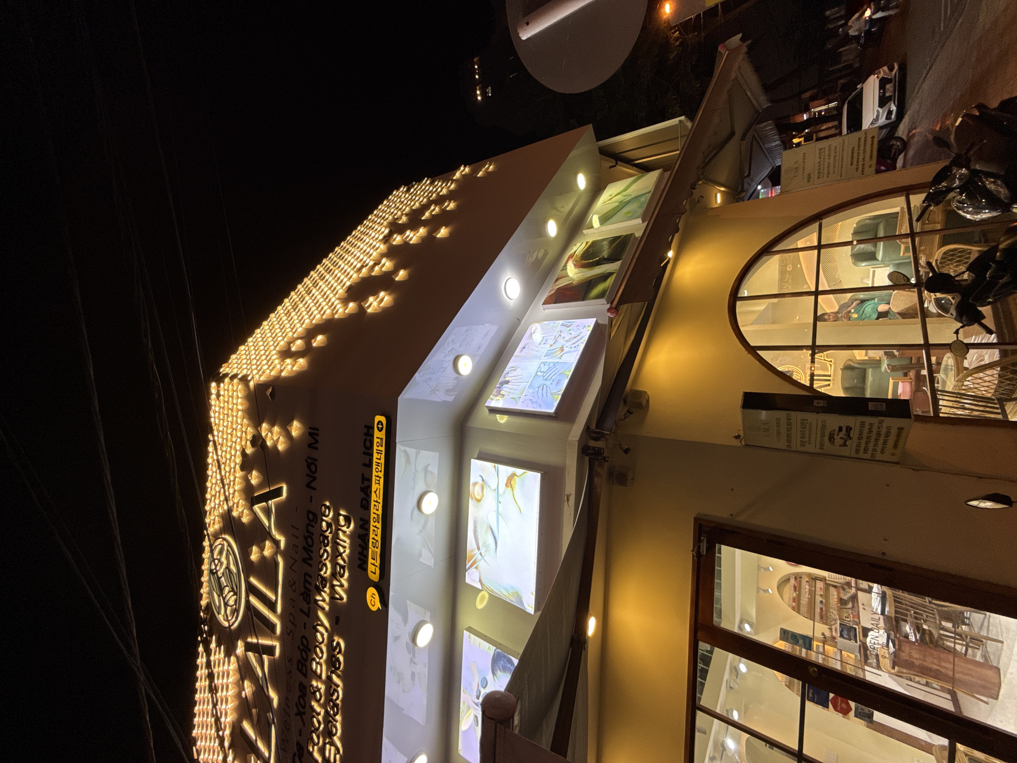 The image shows a brightly lit building at night, likely a spa or salon, on a street in Nha Trang, Vietnam. The building features a prominent sign with illuminated lettering that reads "LAILA" in large font, with smaller text below indicating services like "Wellness Spa & Nail", "Foot & Body Massage", "Eyelashes", and "Waxing". The facade is decorated with numerous small, warm-toned lights.
Below the main sign, there are illuminated panels displaying images that appear to be related to beauty treatments, possibly nail art or spa services. A distinct yellow awning extends from the building over the sidewalk. The building has large windows, revealing the interior which includes seating areas with green chairs and wooden furniture.
On the street in front of the building, there are several parked motorbikes, and a white car is visible further down the road. A person is seen riding a motorbike in the distance. The overall environment suggests an urban setting at night with commercial activity. The sign also includes text in Vietnamese and Korean.