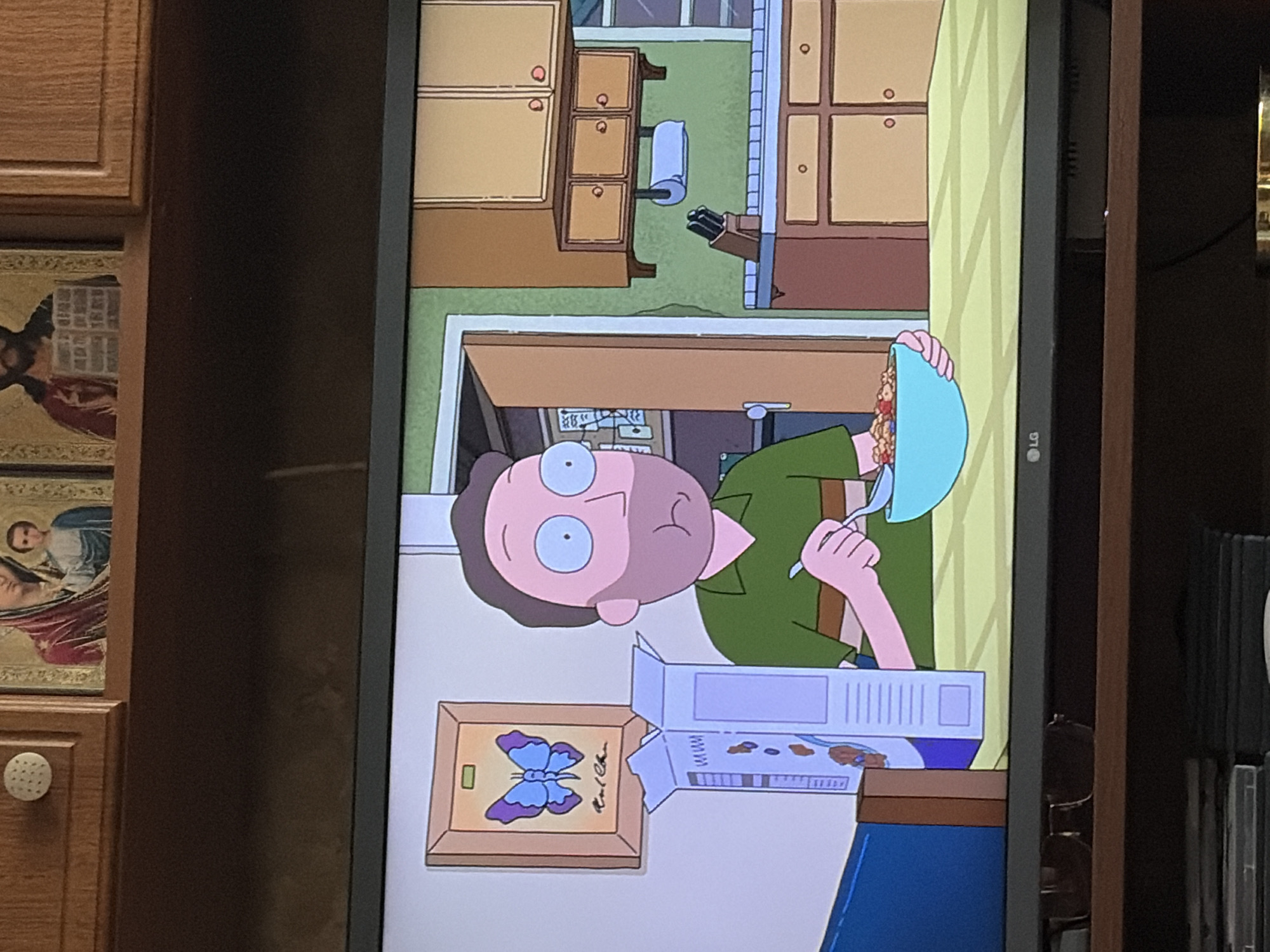 The image shows a scene from the animated television show "Rick and Morty" being displayed on a television screen. The on-screen image depicts a character resembling Jerry Smith sitting at a table, eating cereal. He is wearing a green and brown striped shirt. A blue bowl of cereal and a box of cereal are on the table in front of him. To his left, on the wall, is a framed picture of a blue butterfly. The background shows a kitchen with cabinets and a doorway. The television is placed on a wooden shelf, with the LG logo visible at the bottom of the screen. Above the television, on a wooden cabinet, are two religious icons and a white knob. The overall setting appears to be a domestic interior.