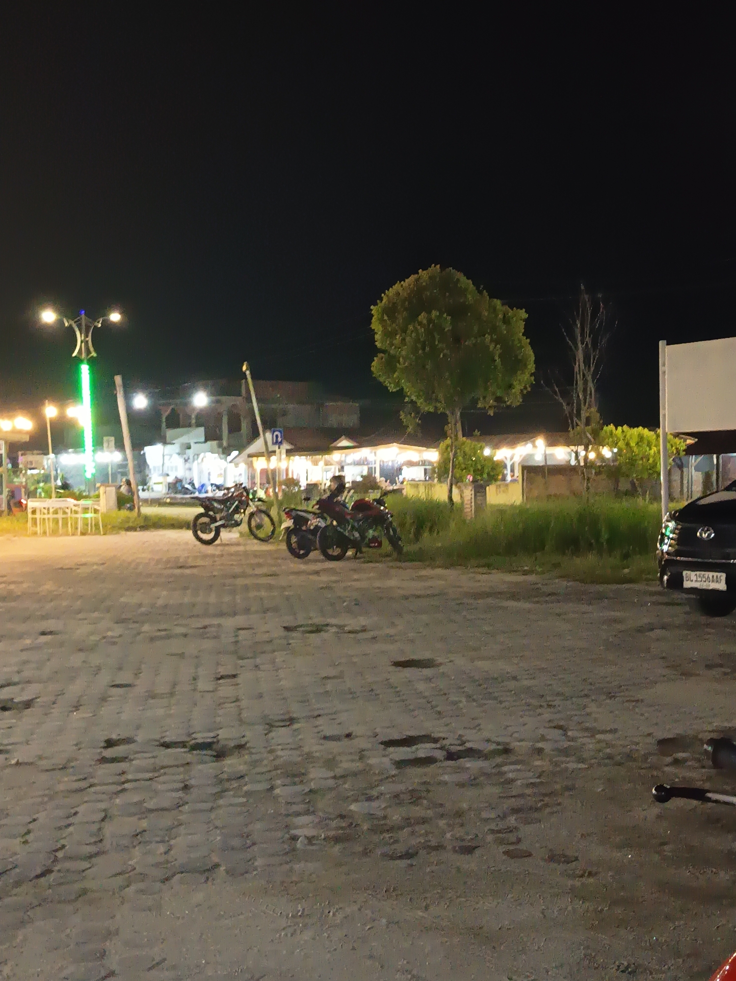 An outdoor nighttime scene in Singkil, Indonesia, depicts a paved area with cobblestone-like patterns. Several motorcycles are parked, including three dirt bikes with black frames and green accents, and one red and black motorcycle. In the background, there are buildings with illuminated awnings and lights, suggesting a commercial or restaurant area. Trees are visible, with one large, bushy tree in the center and a bare, spindly tree to its right. A black car with a license plate reading "BL 1556 AAF" is parked on the right side of the frame. A street lamp with a decorative base and green lights stands on the left. The sky is dark.