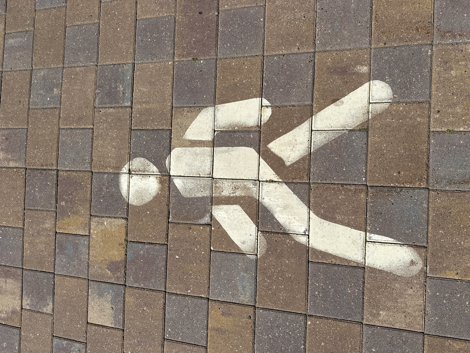 The image shows a pedestrian symbol painted in white on a brick pathway. The pathway is made of rectangular bricks in varying shades of brown and grey, arranged in a staggered pattern. The pedestrian symbol is a stylized representation of a person in motion, with a rounded head, a rectangular torso, and two outstretched arms and legs. The symbol is faded in some areas, suggesting wear and tear from foot traffic. The lighting suggests it is daytime, with shadows cast on the pathway, indicating the sun's position. The image does not contain any specific information or landmarks that would allow for a precise location within Minsk, Belarus.