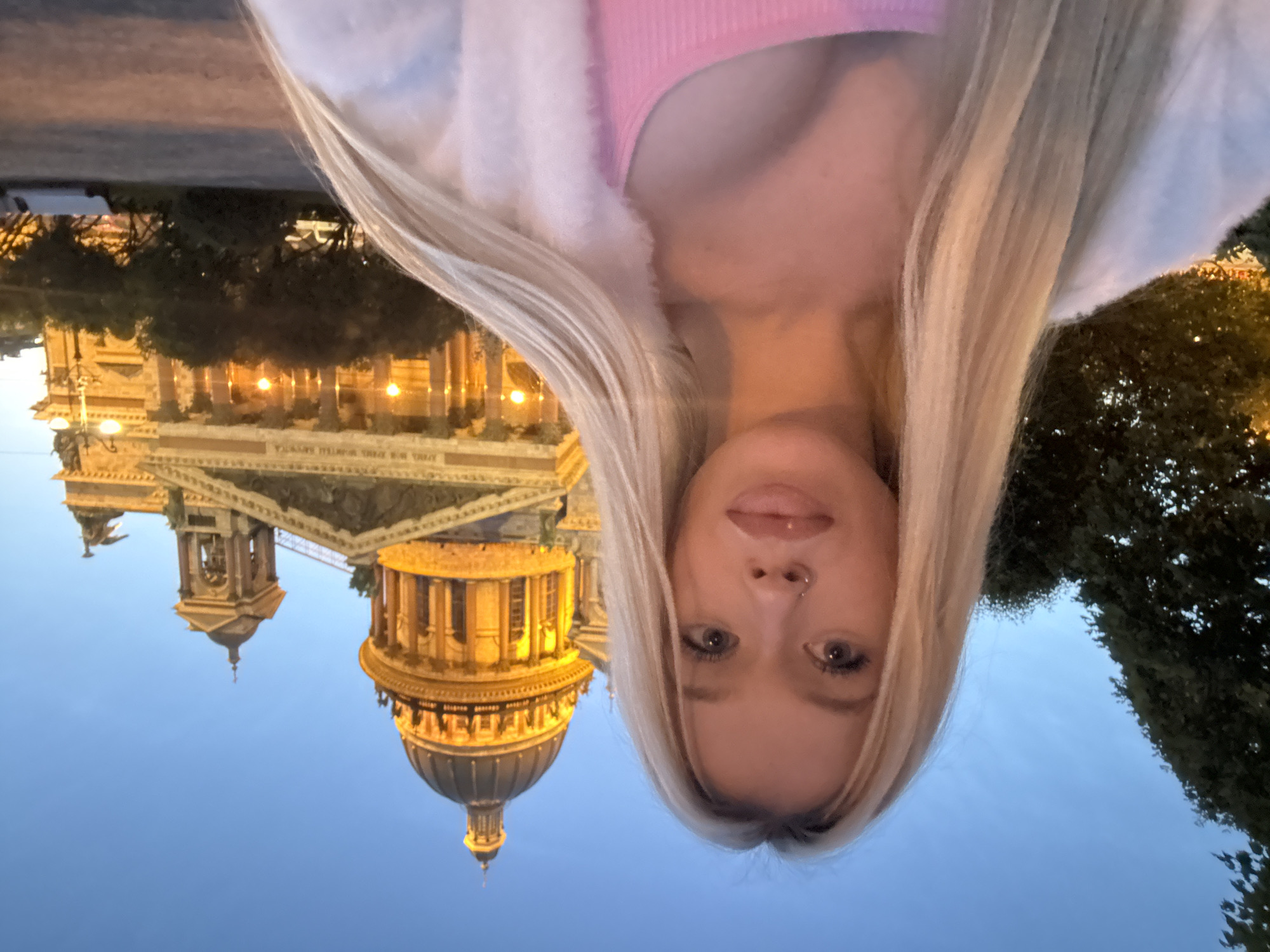 An evening selfie is taken in front of Saint Isaac's Cathedral in Saint Petersburg, Russia. The upper portion of the cathedral, including its dome and facade, is illuminated by warm artificial lights against a deep blue twilight sky. A young woman with long blonde hair and a nose ring is in the foreground, facing the camera. She is wearing a pink ribbed tank top and a fluffy white cardigan. The background features the imposing architecture of the cathedral, trees, and streetlights.