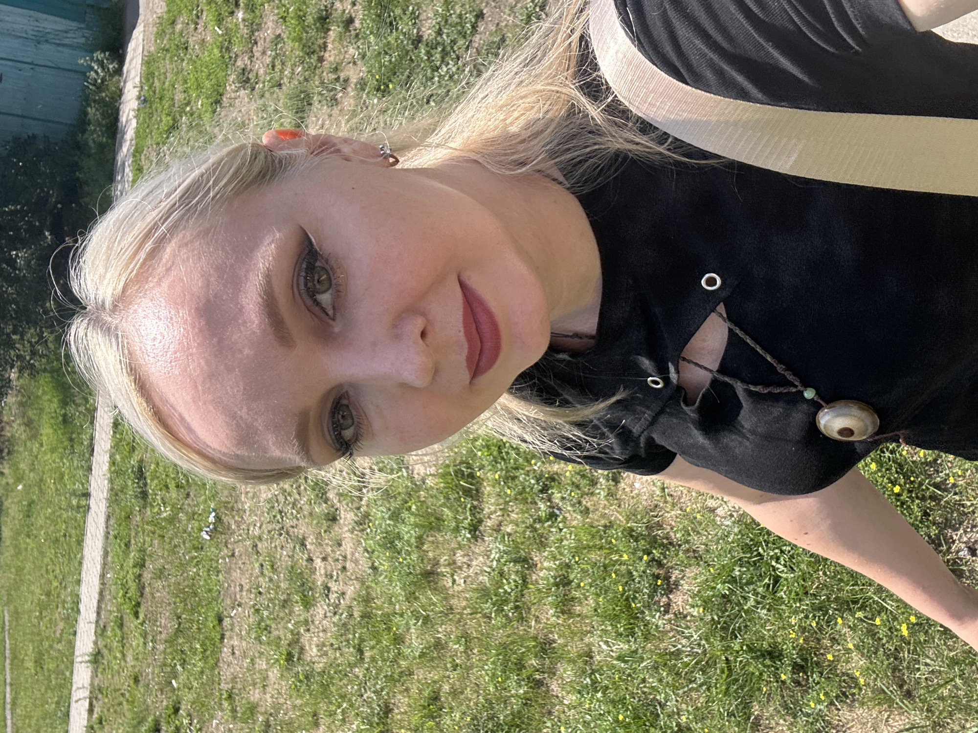 The image shows a close-up, high-angle selfie of a woman with blonde hair and light skin, taken outdoors. She is wearing a black top with a cutout detail and a necklace with a large, circular pendant. She has dramatic eye makeup and reddish lipstick. Her left ear is visible, adorned with a small, heart-shaped earring.
The background consists of a grassy area with small yellow flowers, a paved walkway, and a blurred wooden fence. The lighting suggests it is daytime, likely sunny.
The user requested the location be identified as Komsomolsk-on-Amur, Russia, however, there are no visual cues in the image that definitively place it in that specific city. The environment appears to be a typical suburban or rural outdoor setting.
