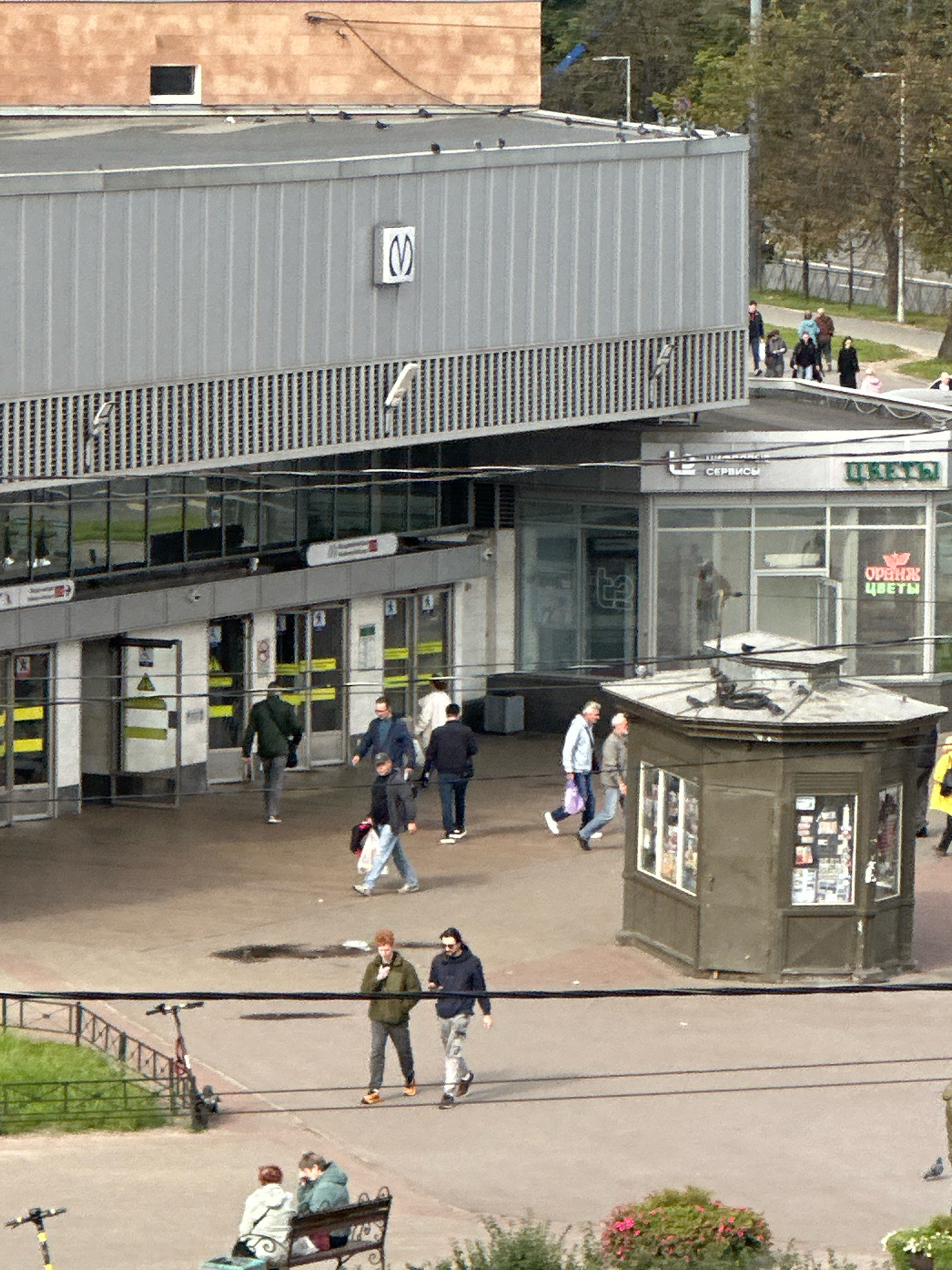 The image captures an overhead view of a busy urban scene in Saint Petersburg, Russia. In the foreground, a paved plaza is populated with pedestrians. To the left, a red electric scooter is parked next to a green fence. Further back, a dark kiosk with a multi-paned roof displays various items on its sides. In the mid-ground, people are walking on the plaza. Behind them is a modern building complex featuring a grey, corrugated metal facade with vertical ribbed detailing. A white square sign with a blue "M" logo is visible on the upper part of the facade. Below this, a glass-fronted structure houses shops, with signage visible, including "ЦВЕТЫ" (Flowers) and "ОРЕНЖ ЦВЕТЫ" (Orange Flowers). The entrance to a metro station is also apparent, with glass doors and yellow and white signage. In the background, trees with autumn foliage suggest the season. The overall atmosphere is one of a bustling public space.