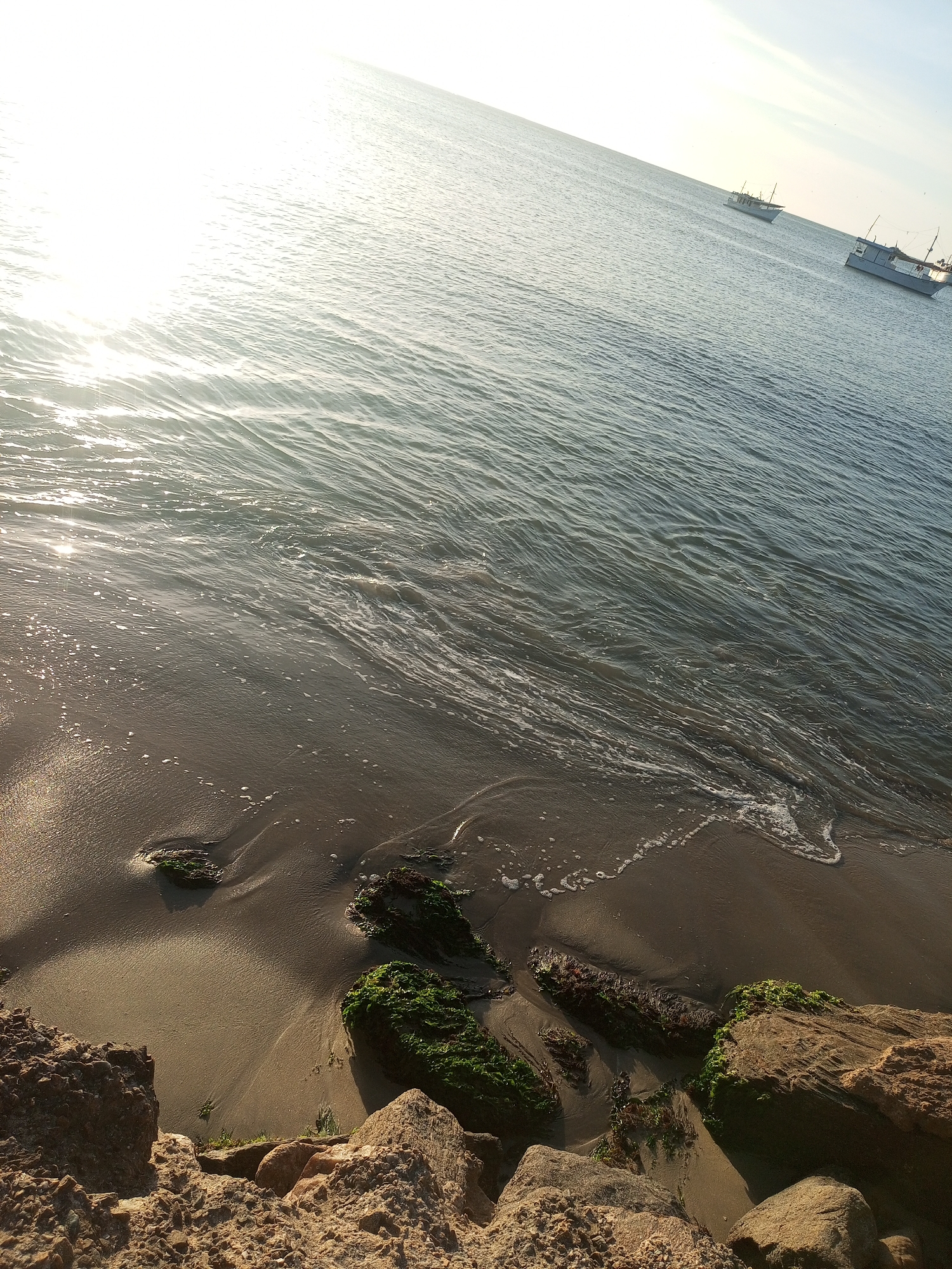 The image depicts a coastal scene in Juan Griego, Venezuela, viewed from a slightly elevated position. The foreground consists of rough, angular brown rocks, some partially covered with bright green algae or moss. Directly beyond the rocks is a sandy shore, wet and textured by gentle waves. Several smaller, dark objects, possibly rocks or wood, are embedded in the sand and also exhibit green algal growth.
The midground is dominated by the calm, grayish-blue ocean water, with small ripples and white foam from breaking waves visible near the shore. Sunlight originates intensely from the upper left, creating a strong, bright reflection across the water's surface in that quadrant. In the upper right background, two white fishing boats are visible on the water, appearing distant with their masts and hulls distinctly silhouetted against the slightly hazy sky. The horizon line is visible, separating the water from a pale, mostly clear sky. No individuals are present in the scene. The overall environment suggests a tranquil marine setting during either sunrise or sunset.