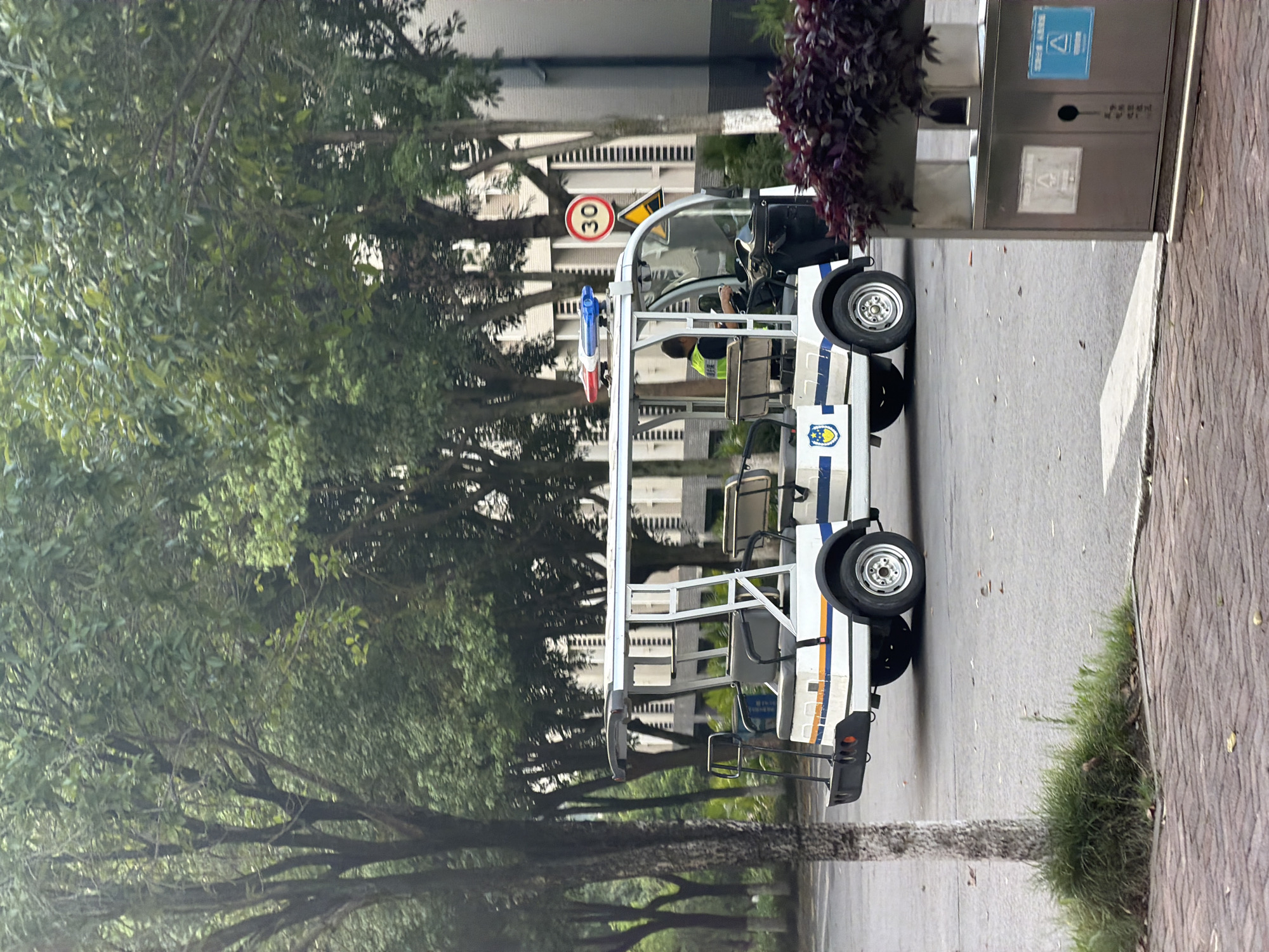 In Jiangjin, China, a white electric utility vehicle with blue and red emergency lights on its roof is parked on an asphalt road lined with trees. The vehicle has a white canopy and appears to be a type of golf cart or shuttle. A person wearing a yellow vest is seated in the driver's seat. A circular red and white speed limit sign displaying "30" is visible on a pole to the right of the vehicle. The environment includes a brick walkway in the foreground, grass, and the facade of a building in the background. A modern metal trash receptacle with signage is situated to the right of the vehicle.