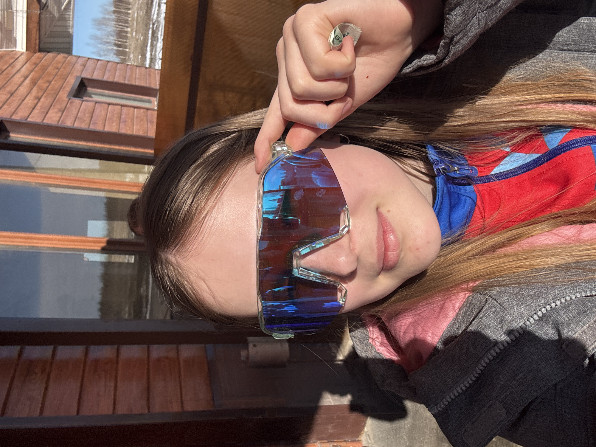 A young woman is outside in daylight near a building with brown horizontal siding and a brown roof, possibly in the vicinity of Gory, Russia. She is wearing a gray zip-up jacket over a red and blue zip-up top, and she is holding a pair of blue-tinted, futuristic-looking sunglasses in front of her face. A folded piece of paper, possibly money, is held near her forehead. The image is oriented sideways.