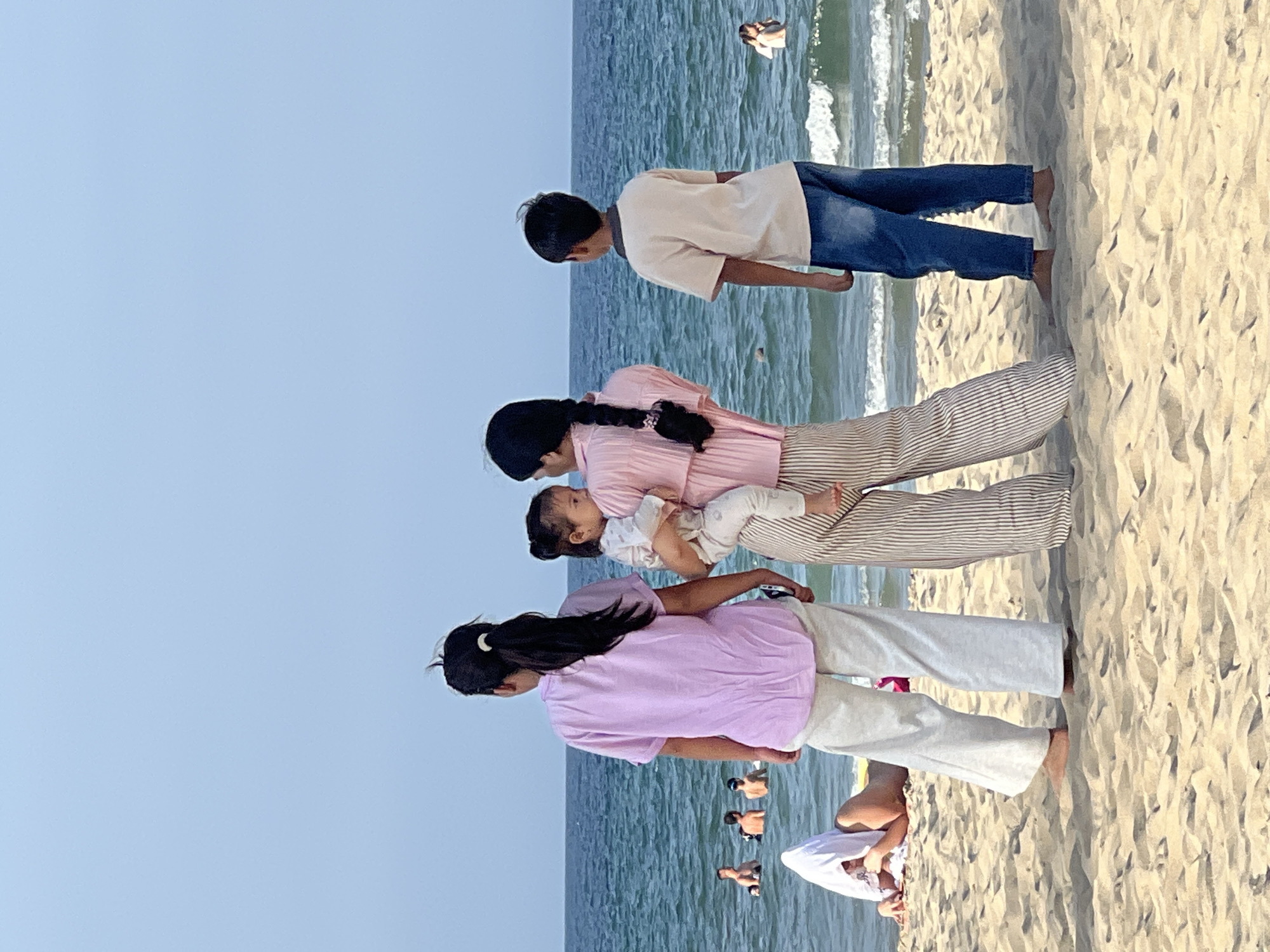 A family is standing on a sandy beach in Da Nang, Vietnam, facing the ocean. In the foreground, from left to right, are a young woman with dark hair in a ponytail, wearing a lavender t-shirt and off-white pants, a baby being held by another person whose face is not visible, wearing a pink shirt and striped pants, and a young boy wearing a light beige t-shirt and blue jeans. The ocean is a vibrant blue with gentle waves. Several other people are visible in the water in the middle ground, appearing to be swimming or wading. In the far background, the horizon is a clear, light blue sky. The sand is light brown and textured with footprints and small debris.
