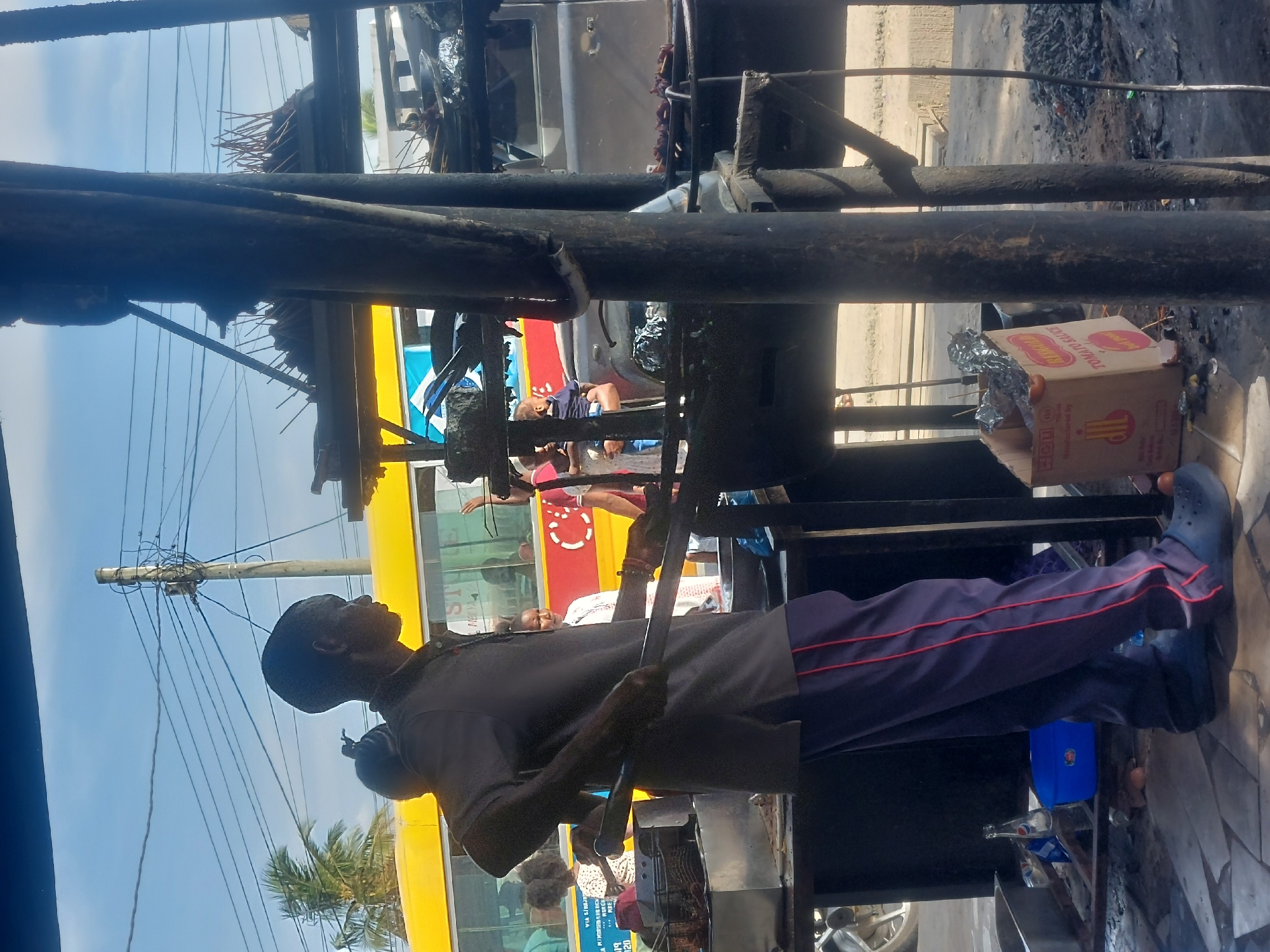 An unedited outdoor photograph shows a street food vendor grilling meat skewers near a bus station in Dar es Salaam, Tanzania. The man is dark-skinned with a black cap, wearing a gray shirt and dark pants with red stripes down the sides, and blue slip-on shoes. He holds a long metal tool in his hands and appears to be tending to a metal grill. The grill is located behind a rusted metal structure, with boxes of "tomato sauce" visible nearby. The ground surrounding the grill is stained and littered with debris. A bright yellow bus full of people is in the background behind the vendor, suggesting a busy urban environment. Overhead power lines are visible in the clear blue sky.