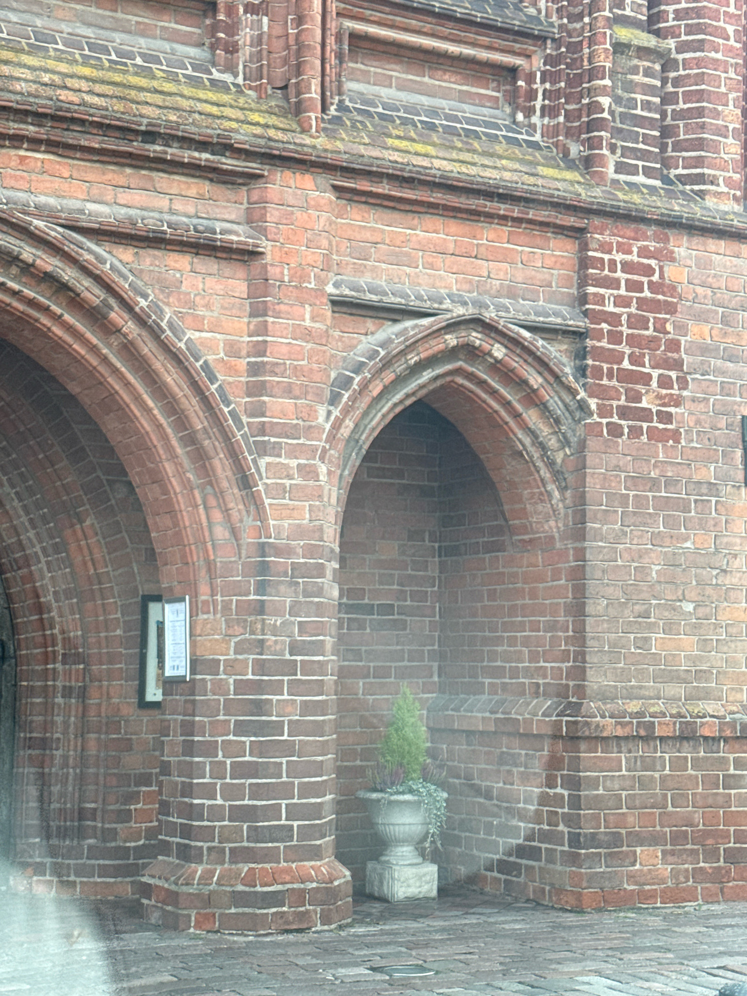 The image shows a close-up view of a brick building with arched entrances. The bricks are a reddish-brown color, and the building has decorative brickwork at the top. In the foreground, a cobblestone path is visible. To the right of the image, a large stone urn contains a small evergreen tree and some trailing plants. A sign is mounted on the wall next to one of the arches. The architectural style suggests it could be a church or another historical building. The overall scene is dimly lit, possibly due to overcast weather or the time of day. This building is located on Pilies Street in Vilnius, Lithuania, and appears to be part of the St. John's Church complex.