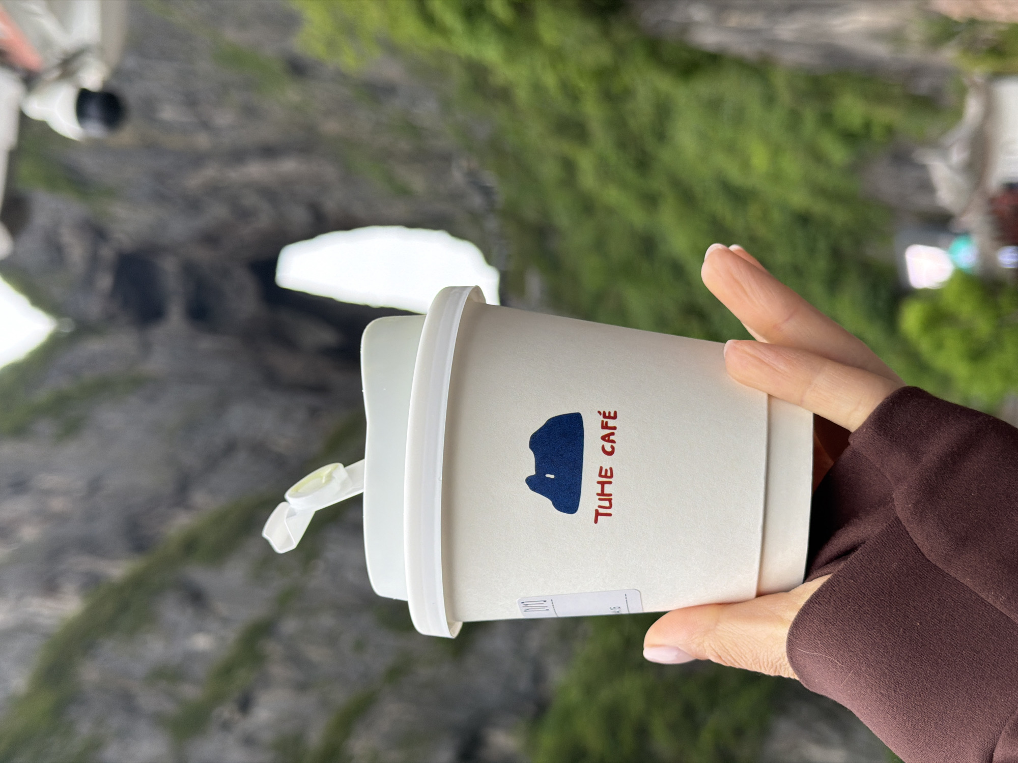 A hand wearing a brown sleeve holds a white coffee cup with the text "TUHE CAFE" and a dark blue graphic on it. A white lid with a straw is on the cup. The background shows a mountainous landscape covered in greenery, with a rocky cliff face and a glimpse of a white structure. The location is likely Zhangjiajie, China, given the unique terrain.