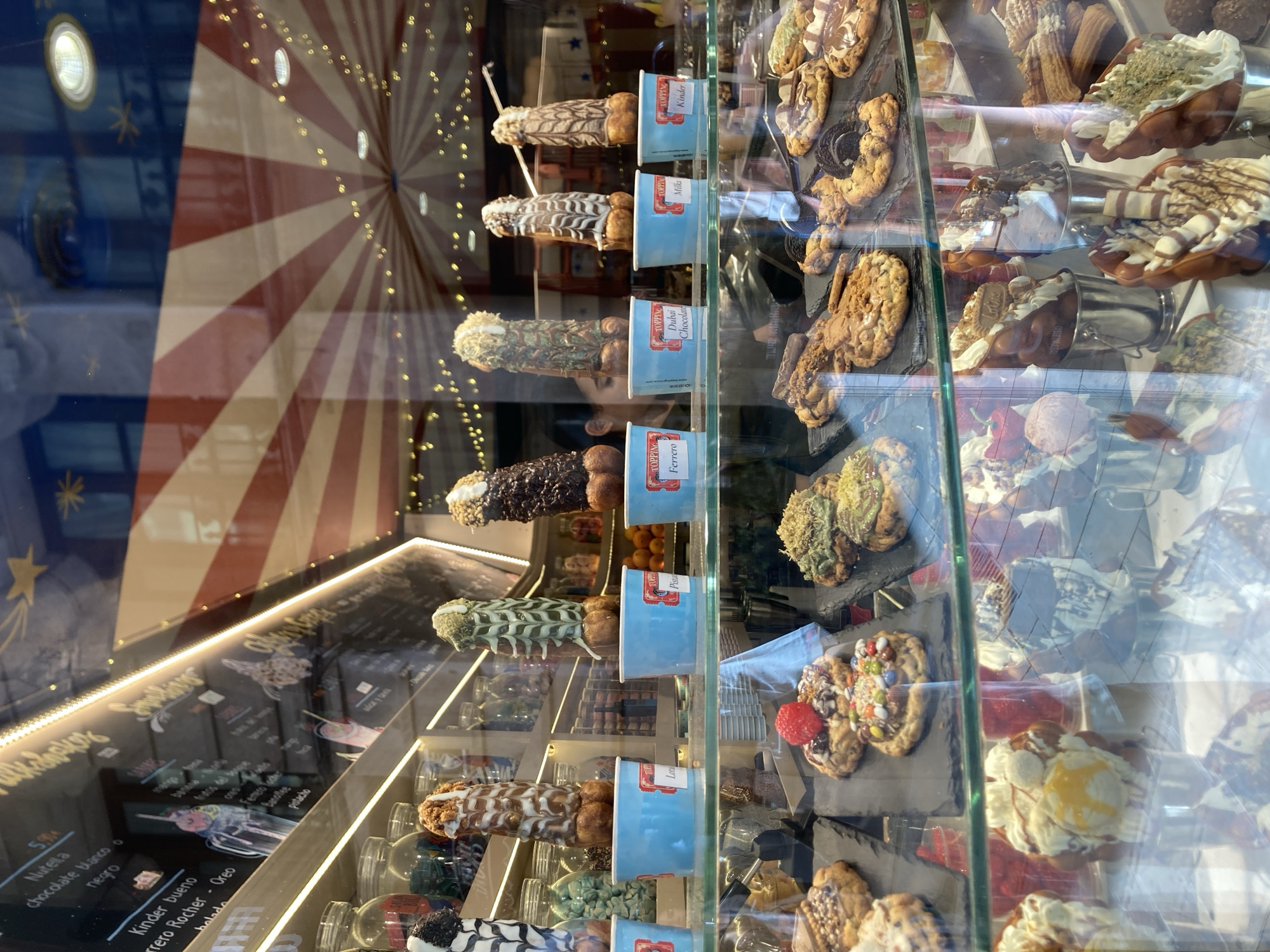 The image shows a shop window displaying various sweets, likely an ice cream or dessert shop, in Barcelona, Spain. The shop has a festive ambiance with a red and white striped awning and string lights visible through the window.
In the foreground, a glass display case holds an assortment of desserts. The top shelf of the display features several penis-shaped waffle cones covered in different toppings like chocolate, white icing, and nuts. Some of these cones are placed in blue cups labeled with flavors such as "Locco," "Ferrero," "Dubai Chocolate," "Milka," and "Kinder." Below these, on a black slate, are cookies topped with chocolate, nuts, and candies, including one with a strawberry. Further down, waffle cones filled with ice cream and toppings are presented in silver buckets.
In the background, through the reflection in the glass, we can see a person's head and shoulders as they appear to be working behind the counter, and some shelves stocked with jars and possibly other ingredients. The overall scene depicts a visually appealing and playful presentation of sweet treats.