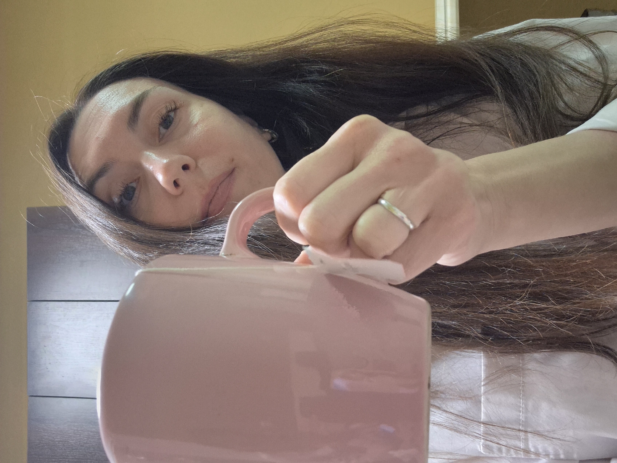 A woman with long brown hair is holding a large pink mug. She is wearing a white shirt and a silver ring on her finger. The background is a wall with wood paneling. The location is Unknown, Unknown.