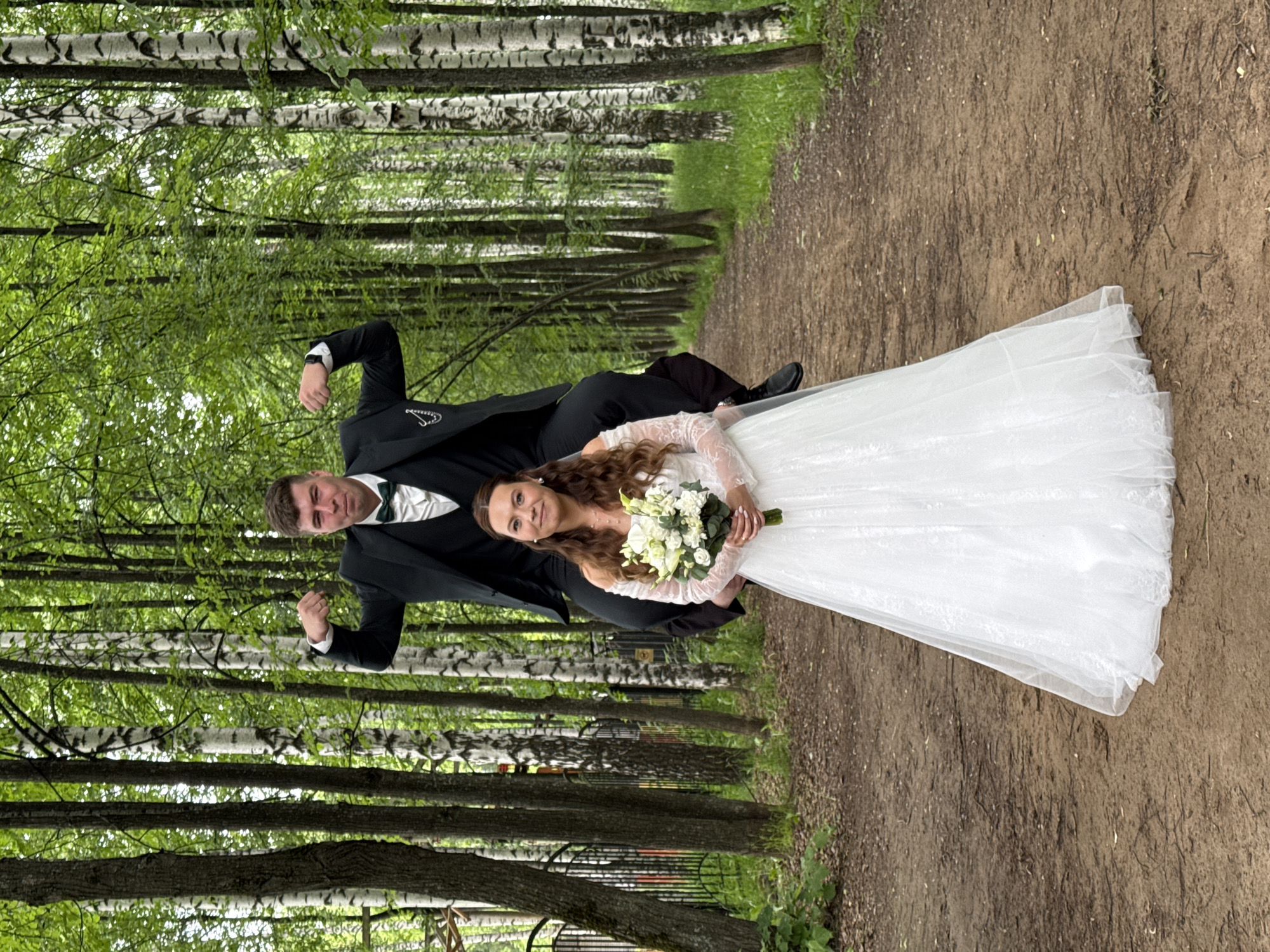 A joyous wedding portrait captures a newlywed couple standing on a dirt path in a tree-lined park or forest, likely in Chaikovsky, Russia. The image is taken during daytime under natural, soft light, suggesting an overcast day or dappled sunlight filtering through the leaves.
The bride, positioned on the right, smiles warmly at the camera, holding a bouquet of white and green flowers, including lilies and other blooms. She is dressed in a traditional white wedding gown featuring elegant long lace sleeves, and her long, curly brown hair is worn down.
To her left, the groom stands in a classic black suit, white shirt, and a dark green bowtie. He strikes a dynamic pose, smiling at the camera with both fists raised in a gesture of triumph or strength. A watch is visible on his left wrist.
The ground beneath them is a reddish-brown dirt path, with sparse green foliage bordering its edges, extending into the distance. The backdrop consists of tall, slender trees, predominantly birch with their distinctive white and black bark, along with other leafy green trees, creating a dense and vibrant green canopy. A black metal fence is subtly visible through the trees in the mid-ground. The overall scene conveys happiness and celebration in a natural, serene setting.
