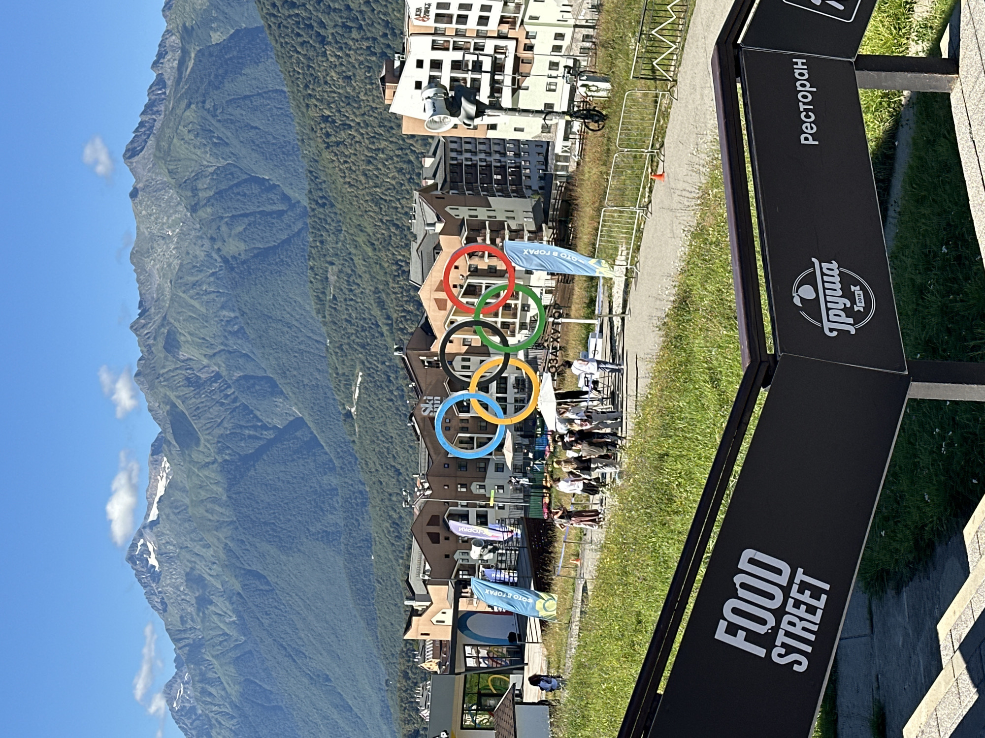 The image depicts an outdoor scene in Estosadok, Russia, likely a resort area, with a prominent Olympic rings monument as a central focal point. The monument, consisting of the iconic interlocking rings in various colors (blue, yellow, red, green, and black), is situated in front of a cluster of multi-story buildings that appear to be hotels or apartment complexes, with an alpine mountain range forming the backdrop.
In the foreground, a sign with the words "FOOD STREET" is visible, indicating a commercial area. Another sign displays "Ресторан" (Restaurant) and a logo for "Груша 2015". A pathway runs in front of the buildings, where a group of people are gathered. Some individuals are standing and observing the Olympic rings, while others appear to be walking or interacting with each other. Several flags with Cyrillic text are planted near the monument.
The environment is sunny with a clear blue sky, and the mountains in the distance show patches of snow, suggesting a high altitude location or a season where snow persists. The surrounding vegetation is green, consisting of grass and trees on the lower slopes of the mountains. The scene conveys a sense of a tourist or recreational destination.