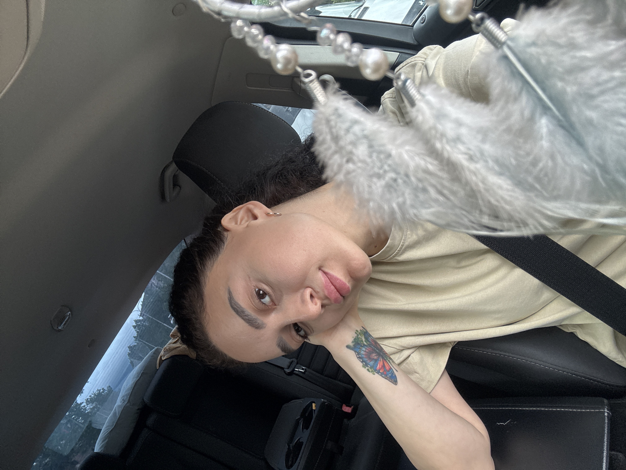 A woman is in the passenger seat of a car. She has a butterfly tattoo on her left wrist and is wearing a beige shirt and a seatbelt. There is a dreamcatcher hanging from the rearview mirror. The location cannot be determined from the image.