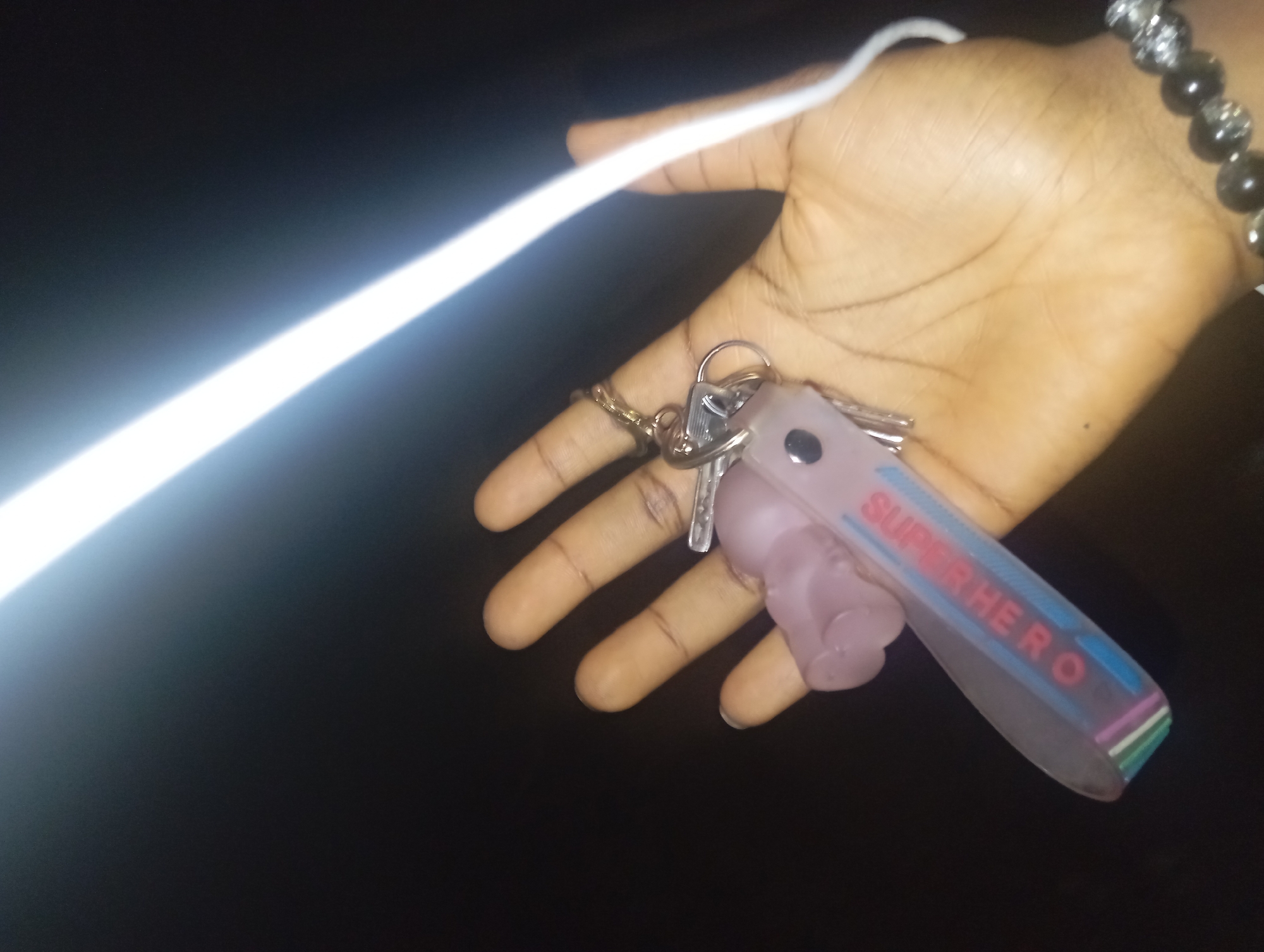 A person's hand is holding a set of keys and a small pink object. The keys are attached to a keychain that has a rectangular, translucent pink tag with "SUPER HERO" printed in blue and red letters. The pink object appears to be a miniature bottle with a cap. The hand is dark-skinned with visible fingerprints. The background is dark and out of focus, with a bright white light source diagonally across the upper portion of the image. A bracelet with dark beads and silver accents is visible on the person's wrist. The scene is likely indoors or outdoors at night, given the lighting conditions. No specific location within a city can be determined from the image.